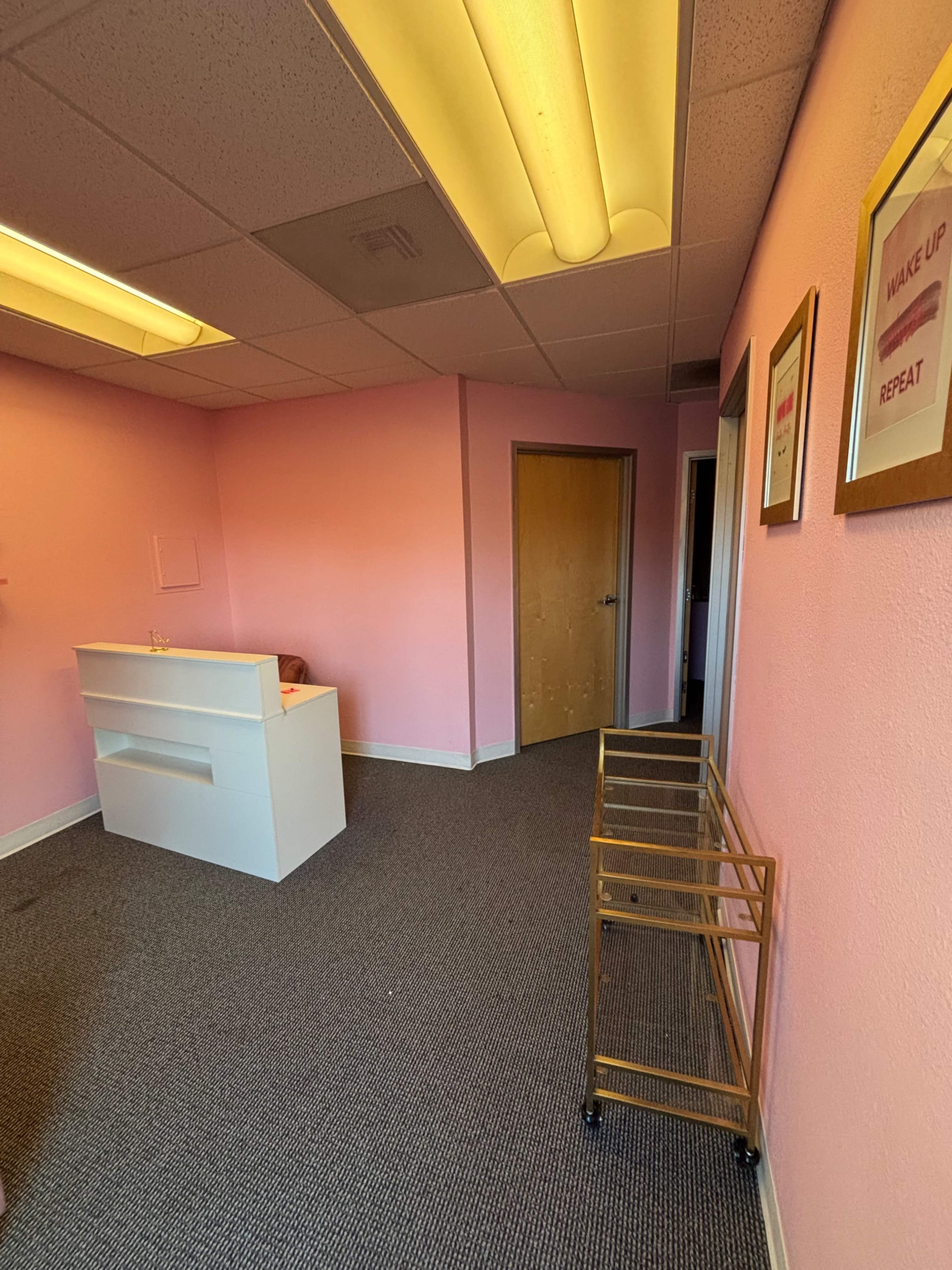 The image shows a pink-walled room with a white reception desk, a rolling cart, and framed motivational posters on the walls.