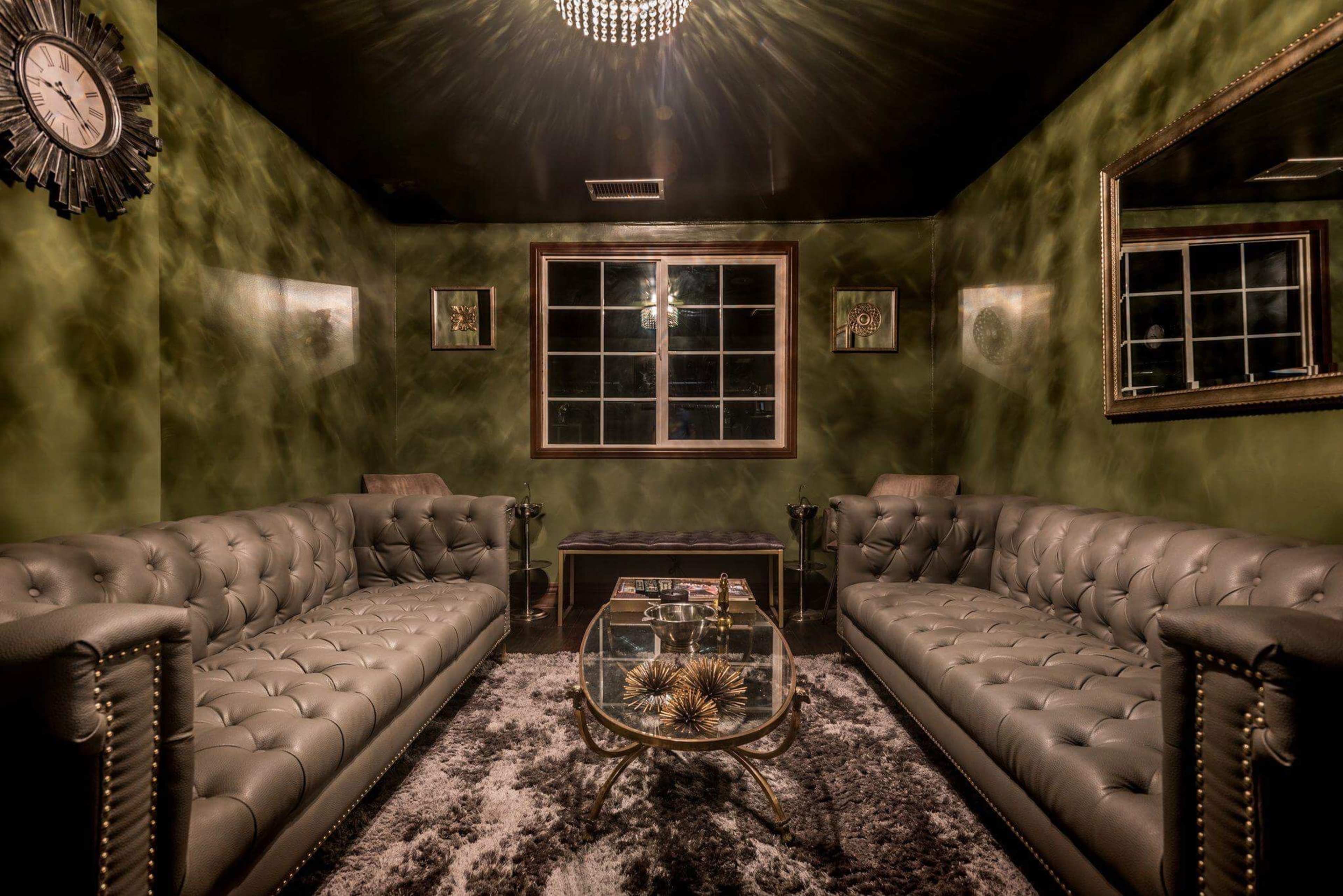 The room features two tufted leather sofas, a glass coffee table, and a large window, all accented by dark green walls and decorative mirrors.
