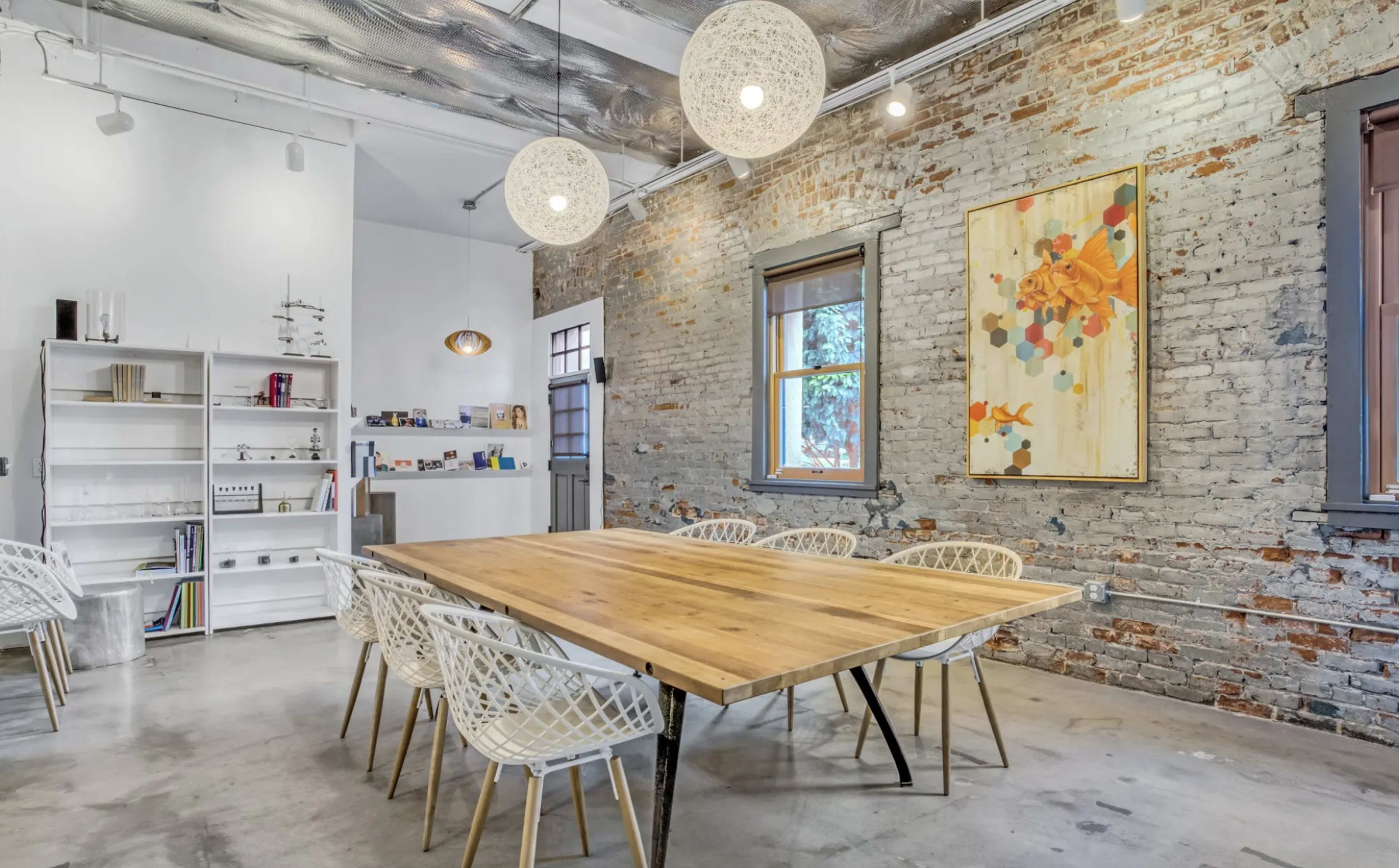 The image shows a modern meeting room featuring a large wooden table surrounded by white chairs, exposed brick walls, and pendant lighting.