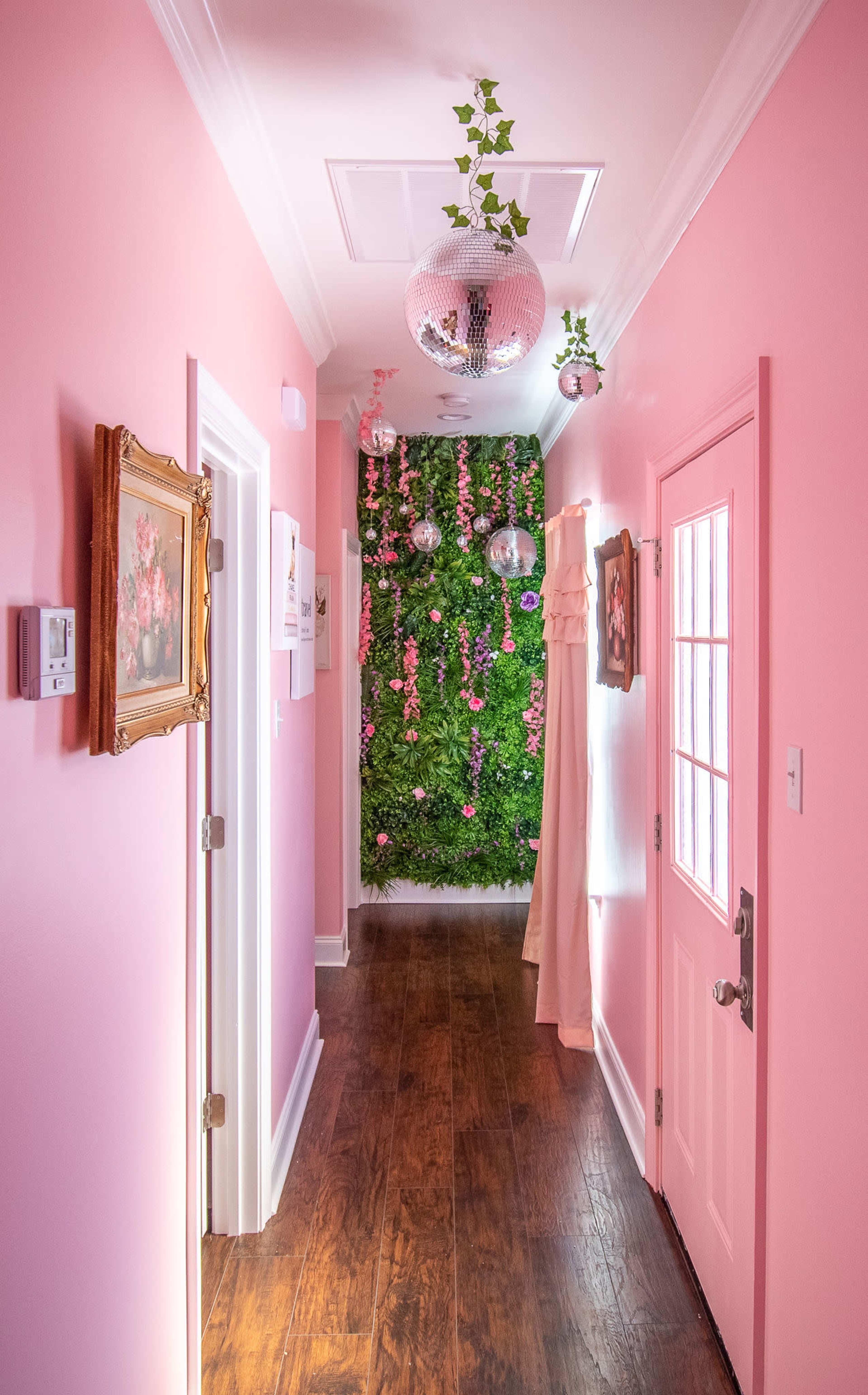 A pink hallway features a green vertical garden along one wall, adorned with hanging plants and disco balls, leading to a brightly colored door.