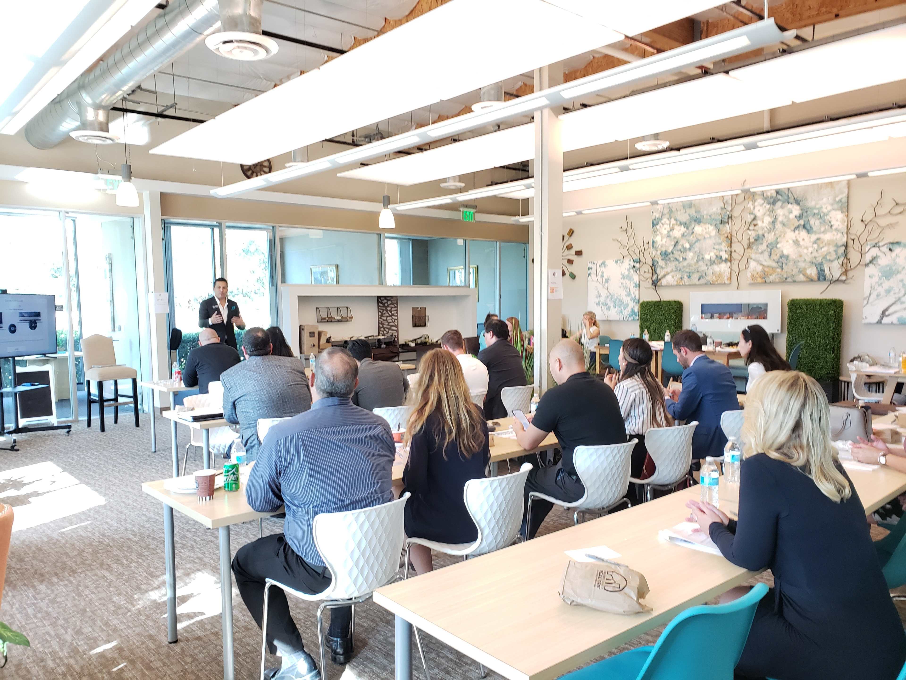 A speaker presents to an audience seated at tables in a modern conference room.