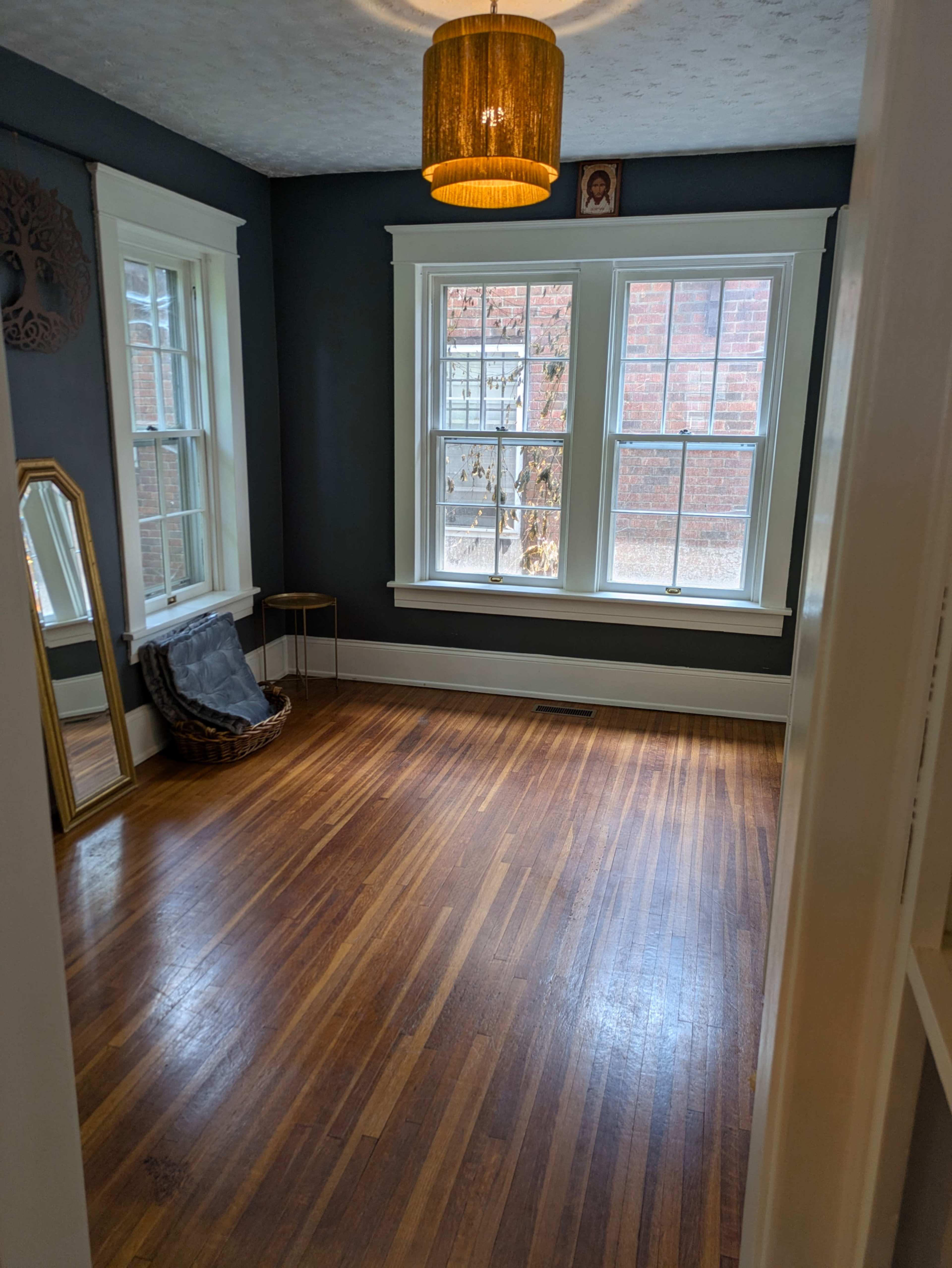 The image shows a well-lit empty room with hardwood floors, two large windows, a wall-mounted picture, and a gold-colored lamp hanging from the ceiling.