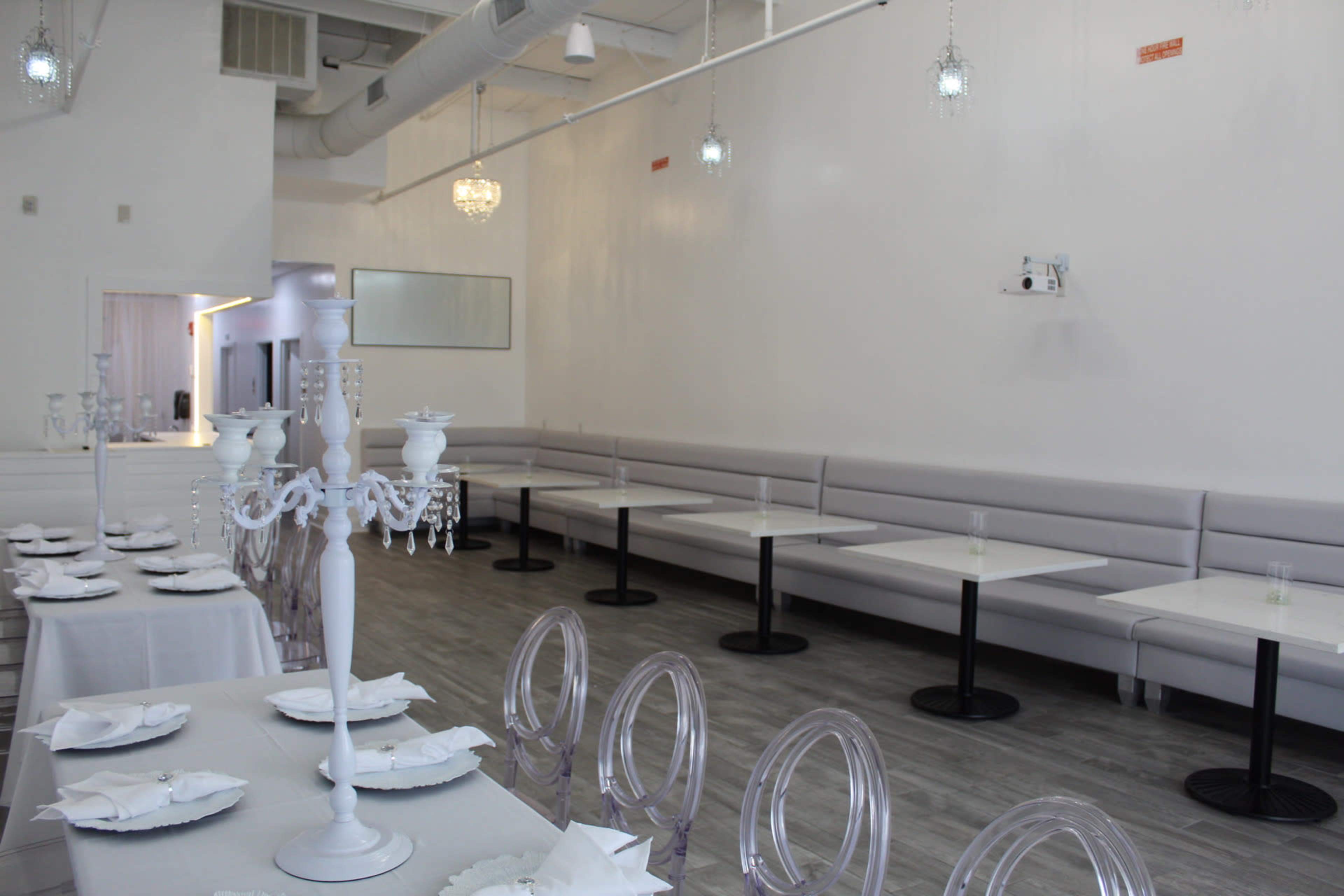 The image shows a modern restaurant interior with white walls, gray flooring, several tables with white tablecloths, and transparent chairs arranged along a row of booths.