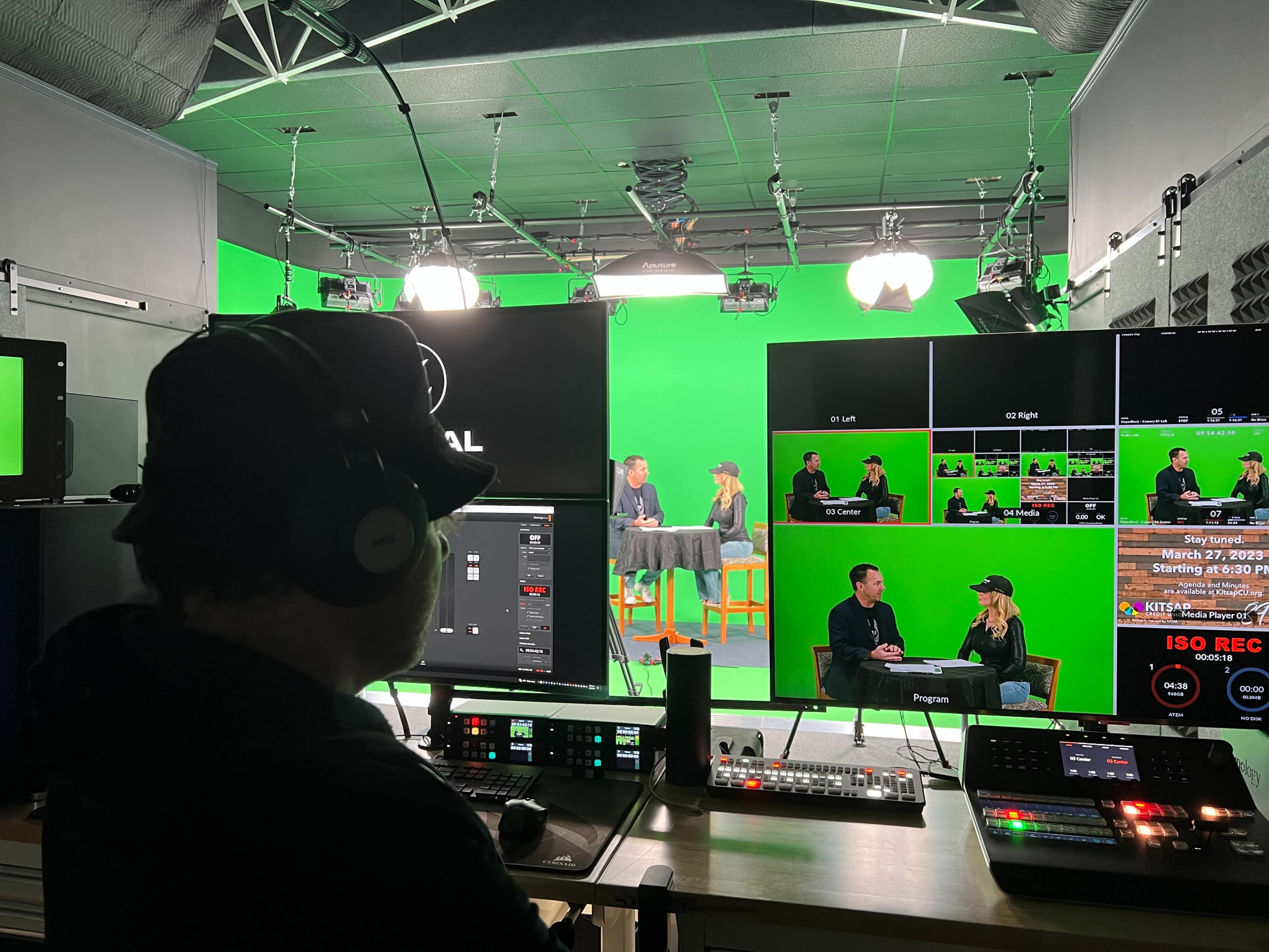 A person operates a video production setup in a studio with green screens and multiple monitors displaying a live broadcast.