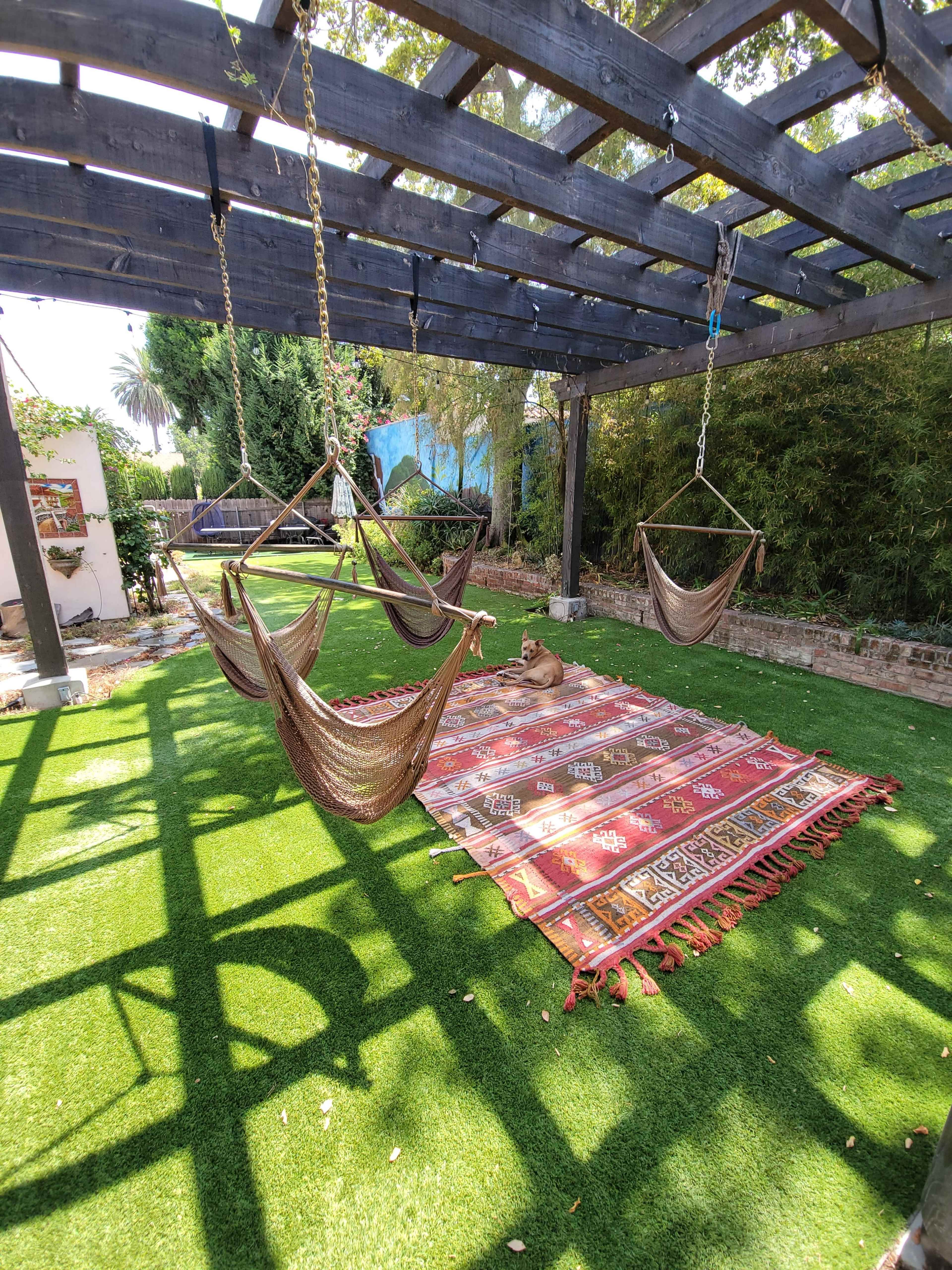 A shaded outdoor space features three hanging hammocks above a large decorative rug on artificial grass.