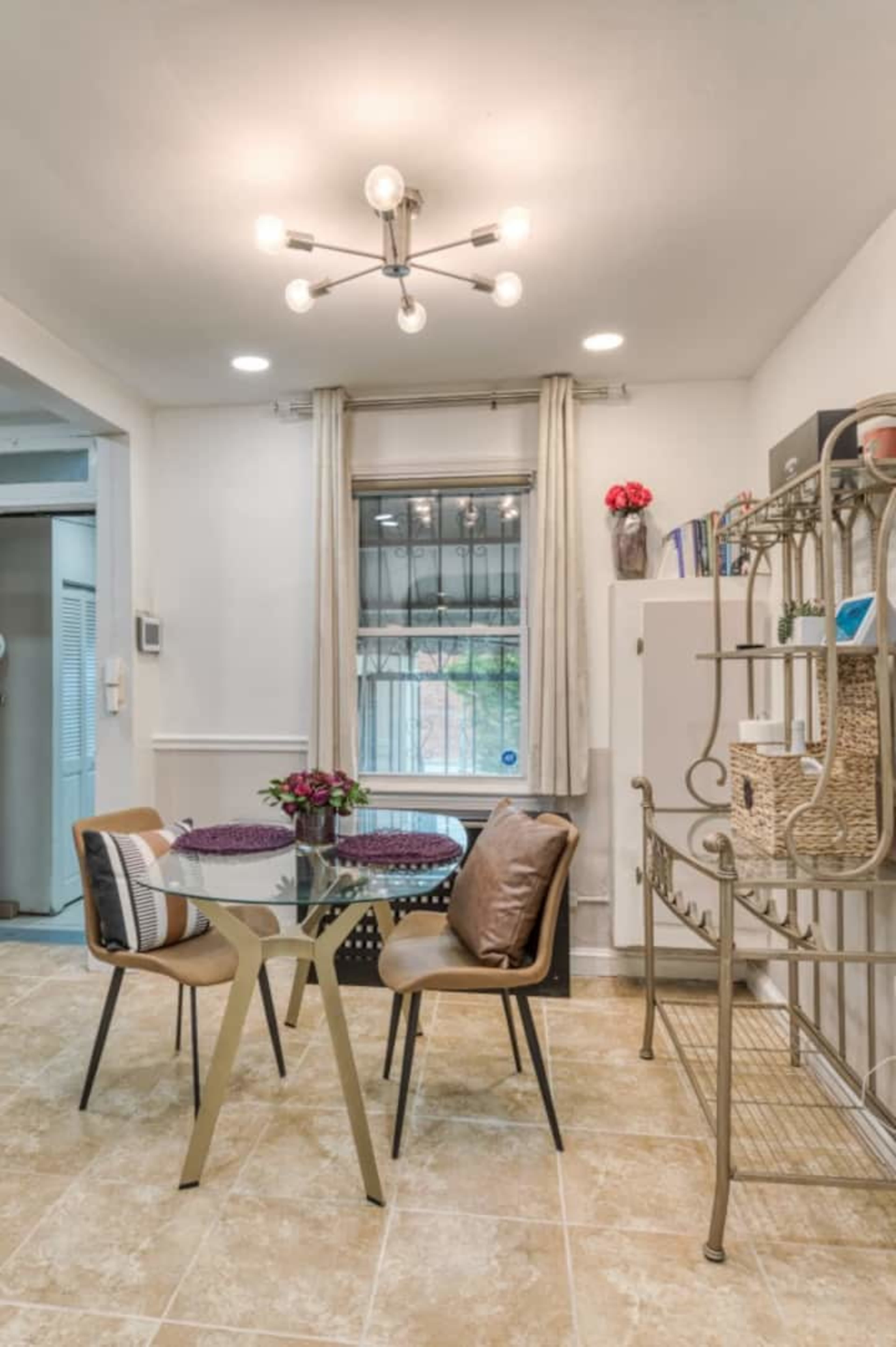 A small dining area features a round glass table with two chairs, a window with curtains, and a metal shelving unit against the wall.