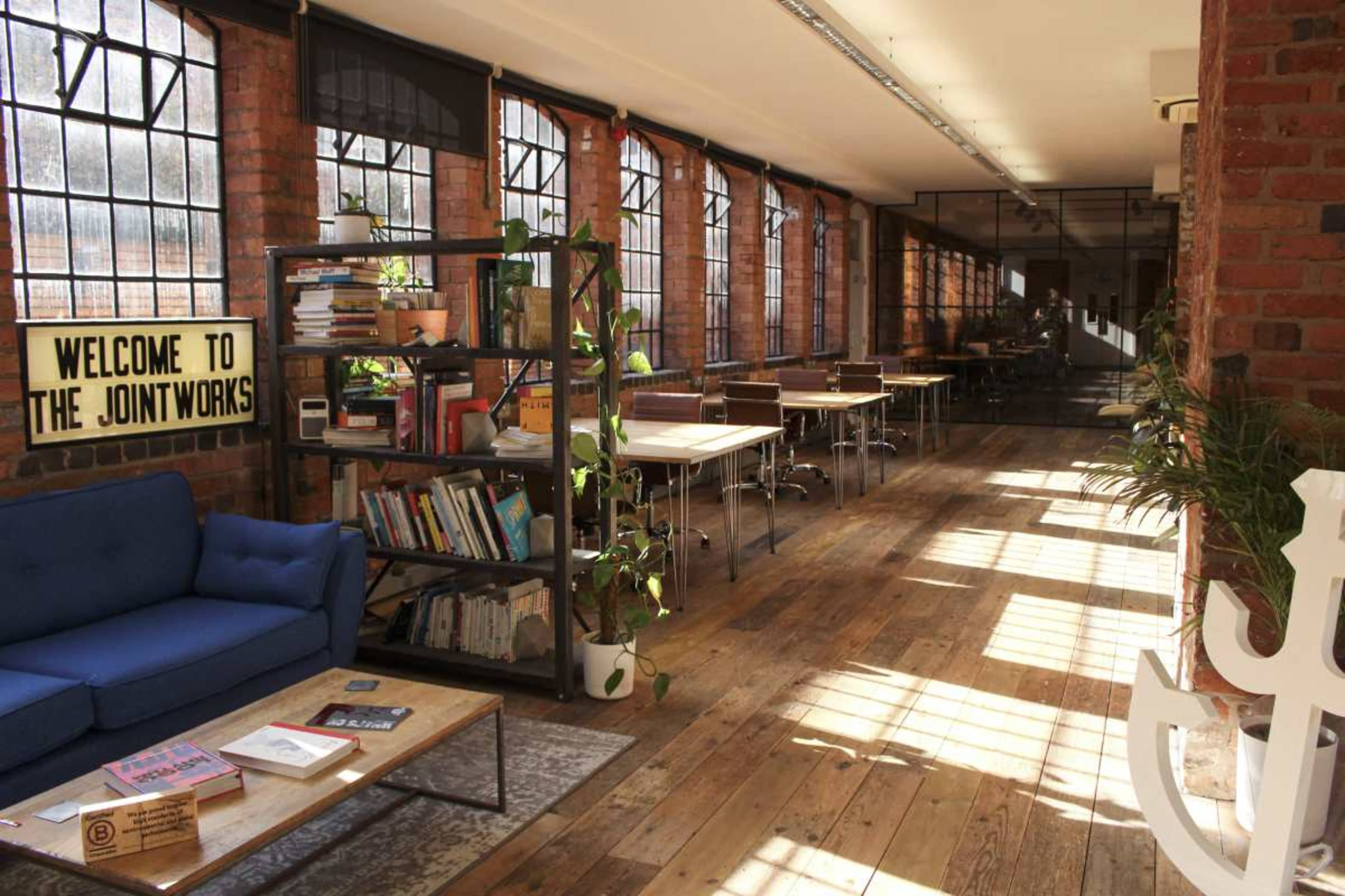 The image shows a bright, spacious coworking area with large windows, wooden flooring, a blue sofa, book shelves, and several tables for work.