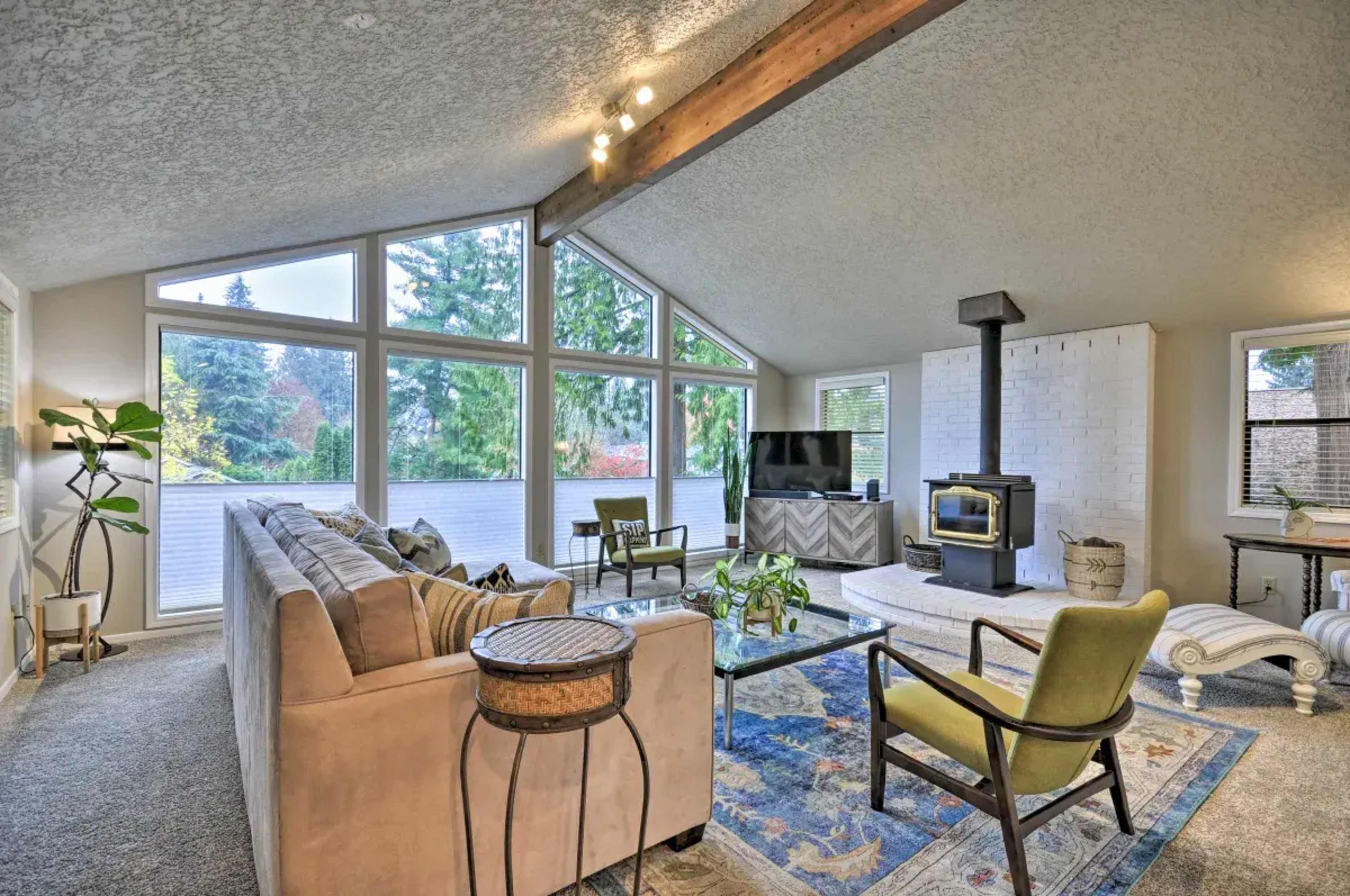 A cozy living room with a high ceiling, large windows, a wood stove, and modern furniture arranged around a patterned rug.