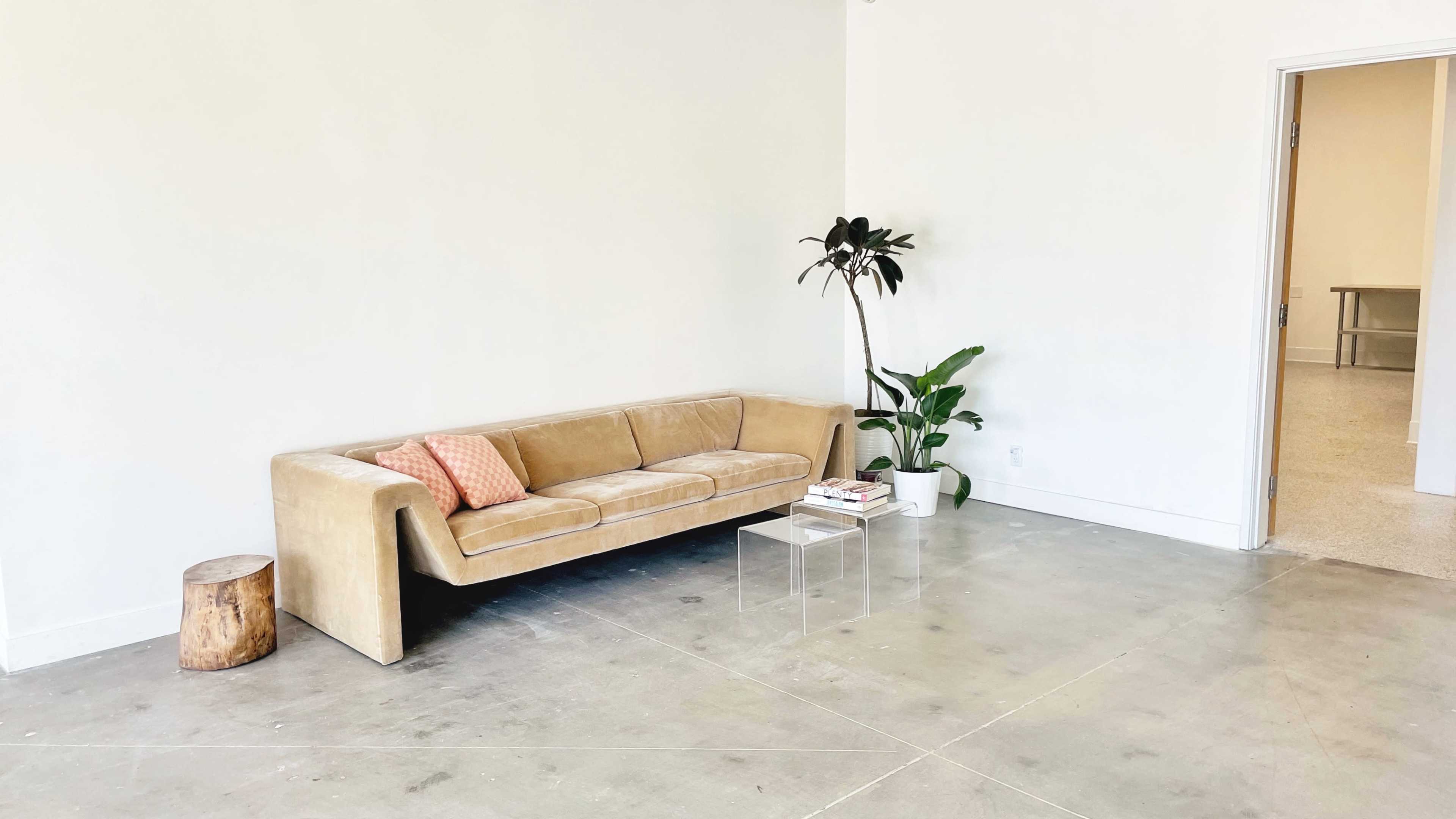 A minimalist living area featuring a light-colored sofa with an accent pillow, a small wooden stool, a potted plant, and a clear acrylic coffee table on a concrete floor.