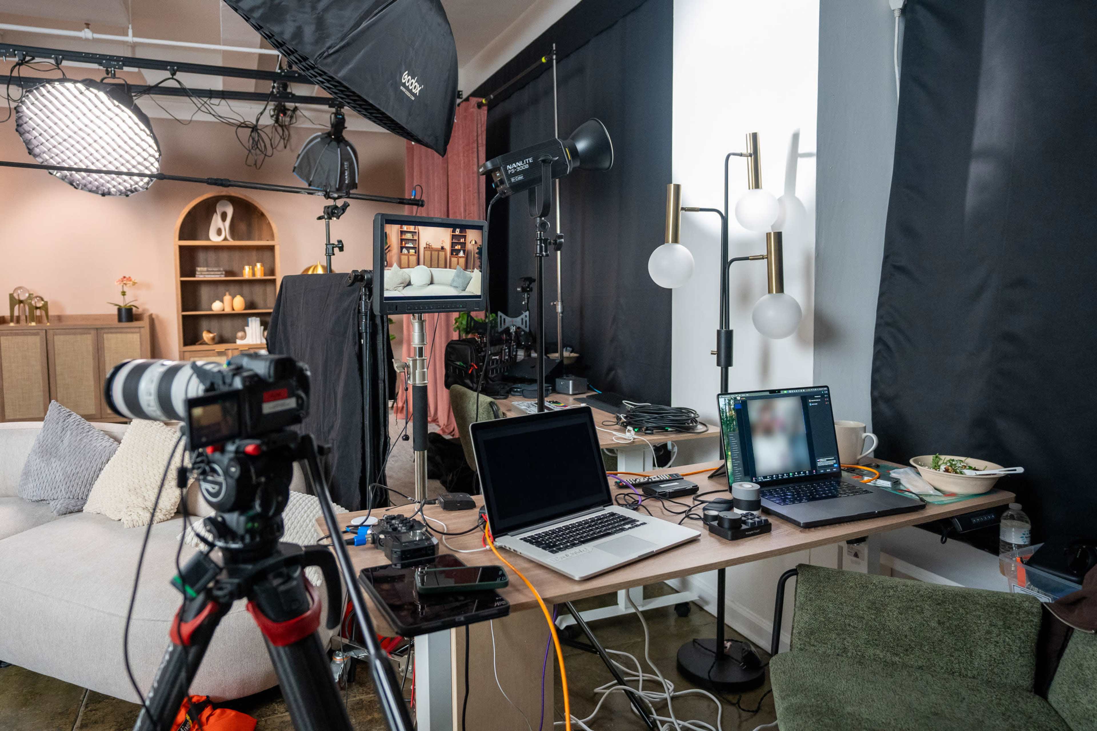 A cluttered workspace features a camera setup, laptops, lighting equipment, and various cables, alongside a small table with food and drink.