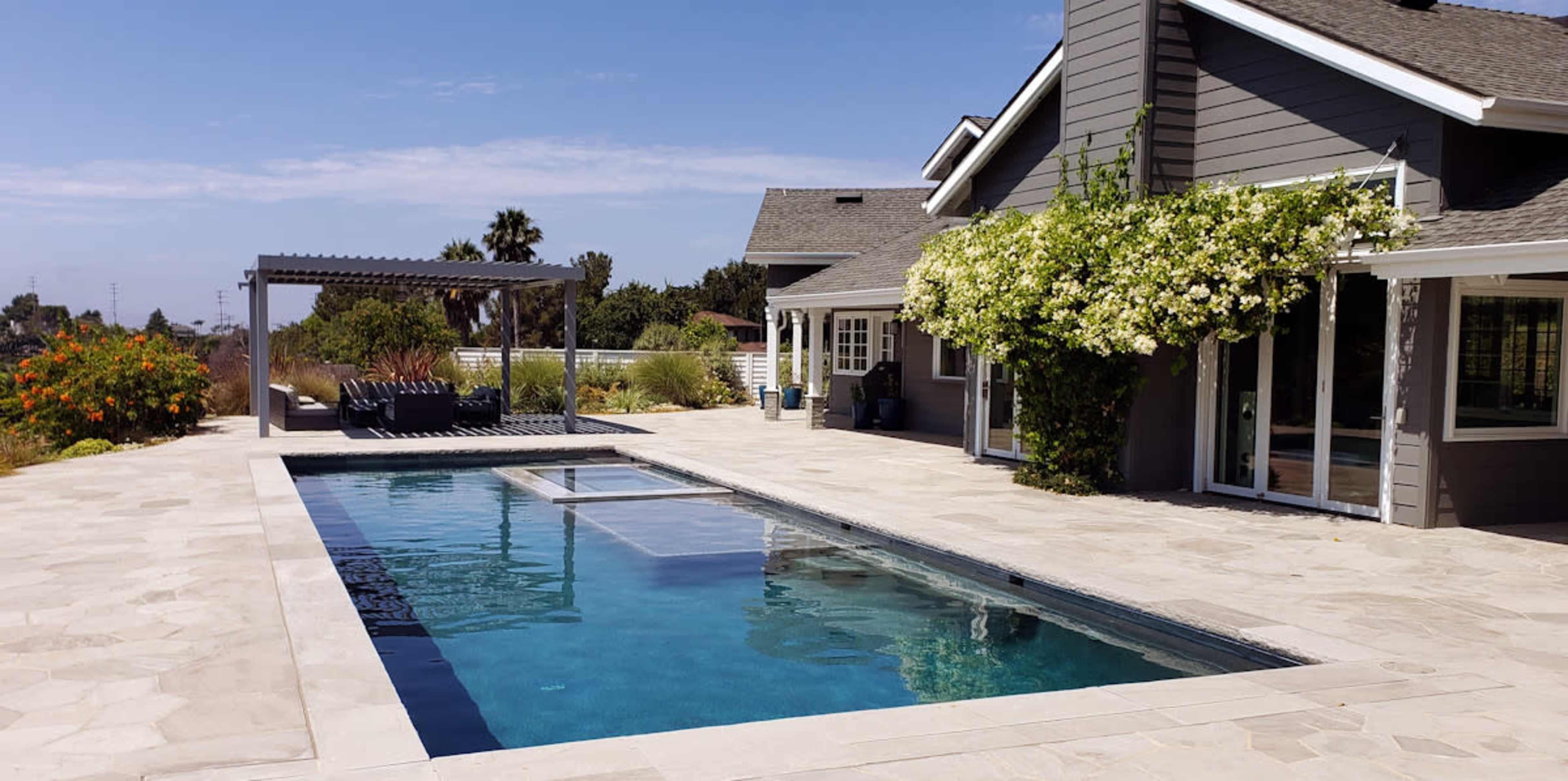 A rectangular swimming pool surrounded by a stone patio and a house with a grey exterior and flowering plants.