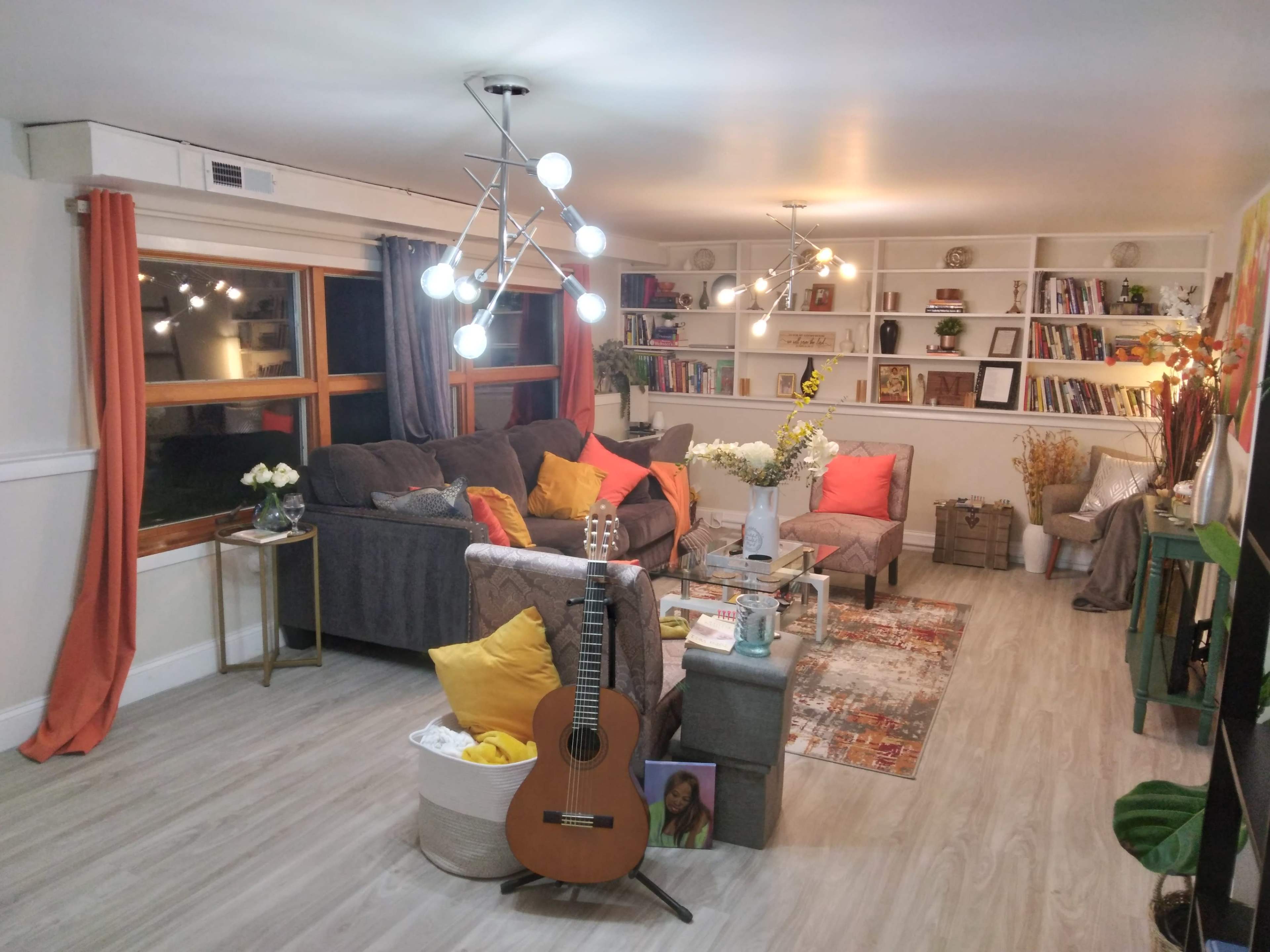 The cozy living room features a gray sofa, colorful cushions, bookshelves filled with books and decor, and a guitar placed against a coffee table.