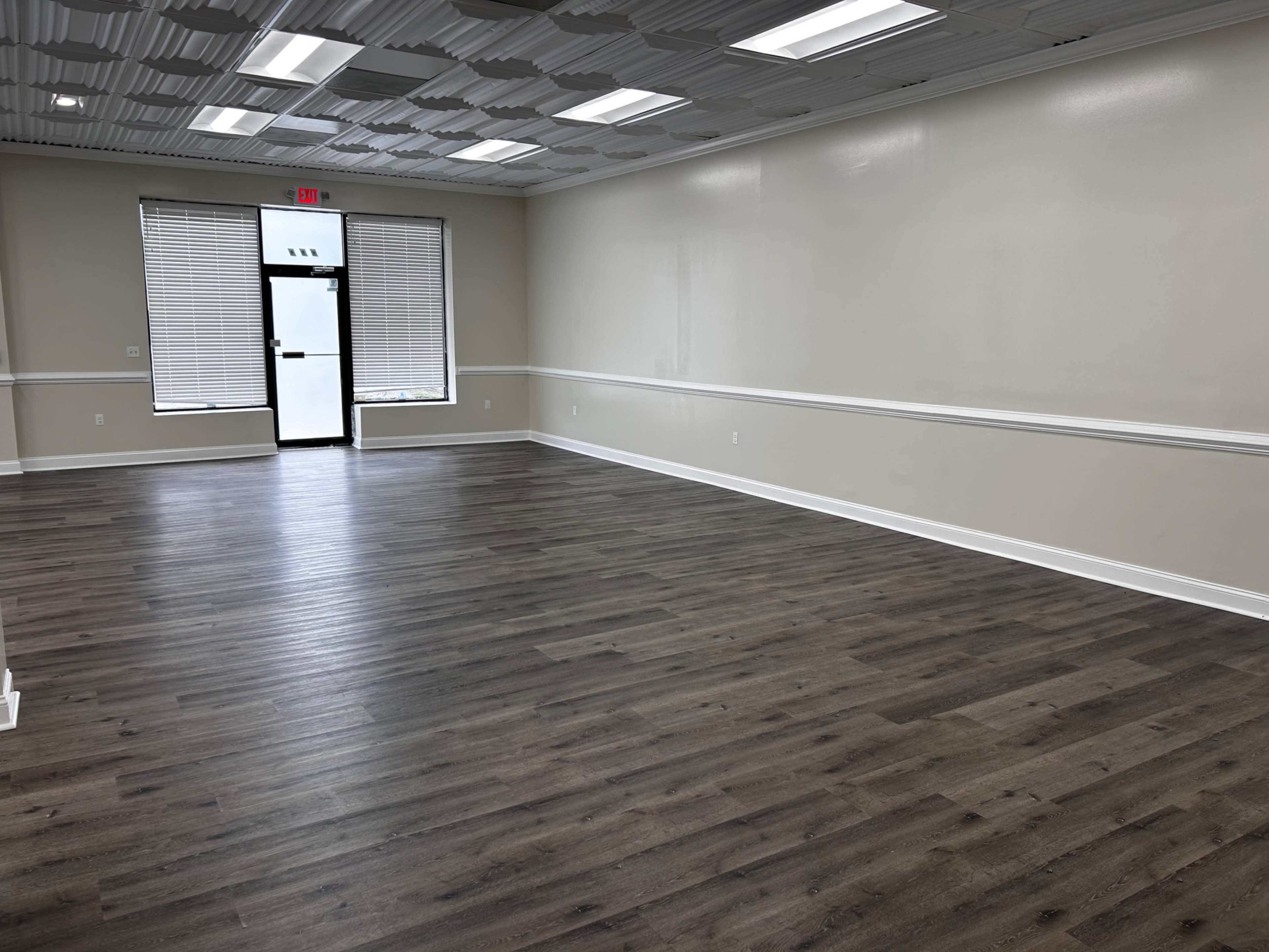 The image shows a vacant interior space with a polished wooden floor, white walls, and a large entrance door with windows.