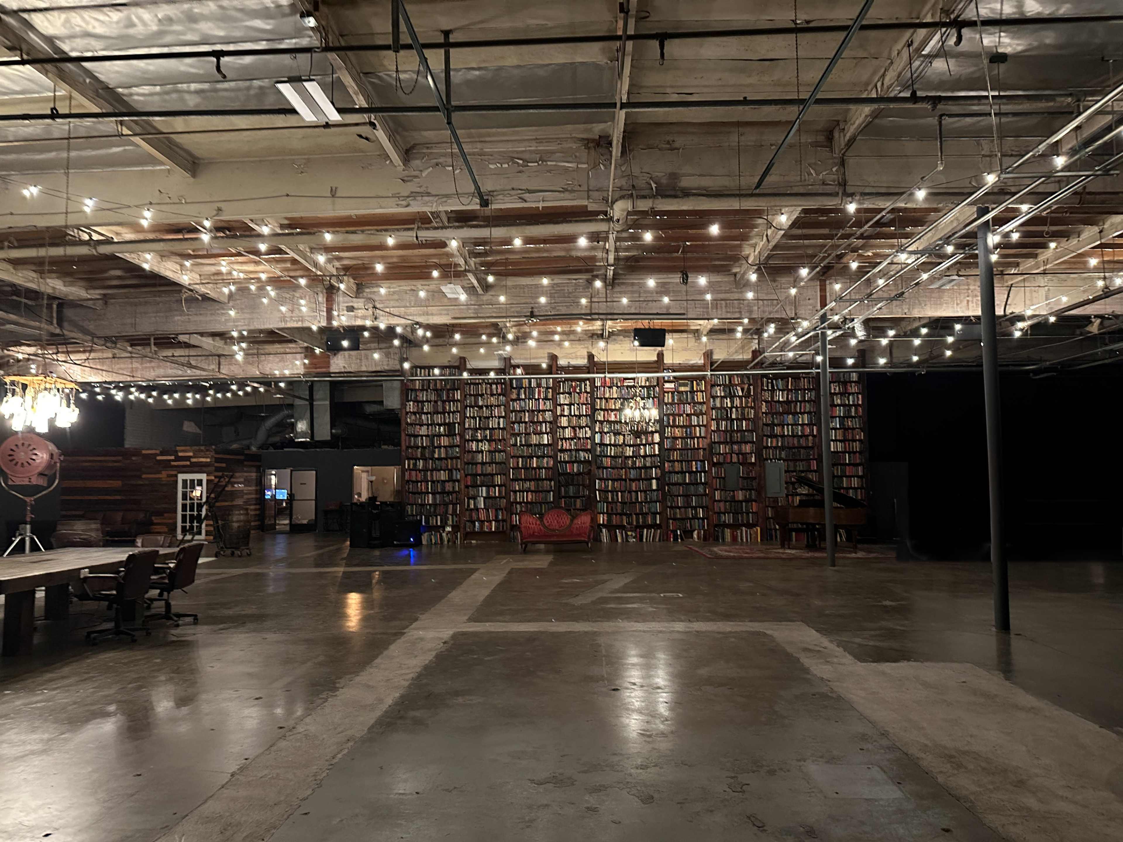 A spacious, industrial-style room with a large bookshelf wall, scattered lights overhead, a red sofa, and a long table with chairs.