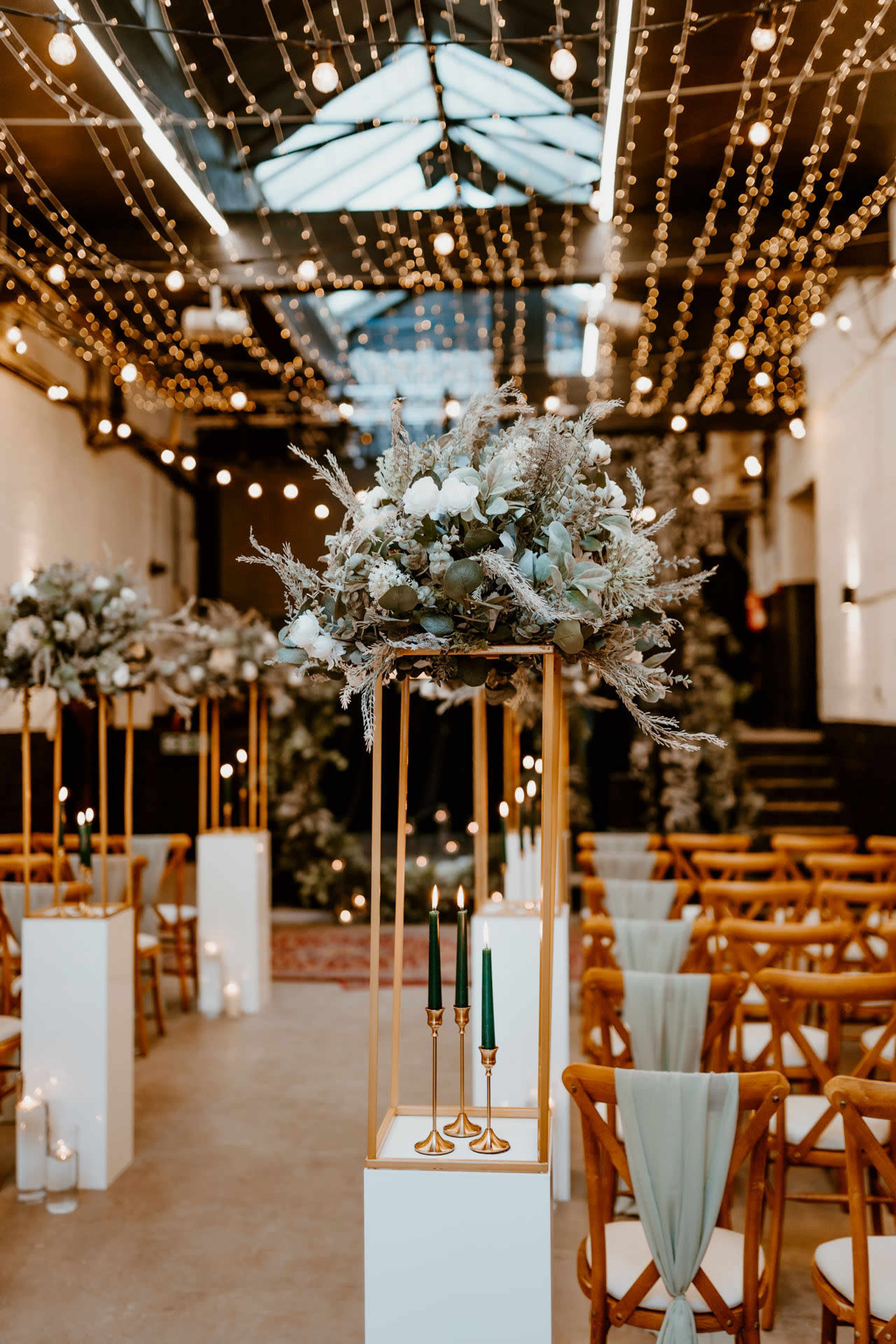 A wedding ceremony setup features tall floral arrangements on pedestals, with green candles and seating arranged in front, all illuminated by string lights.