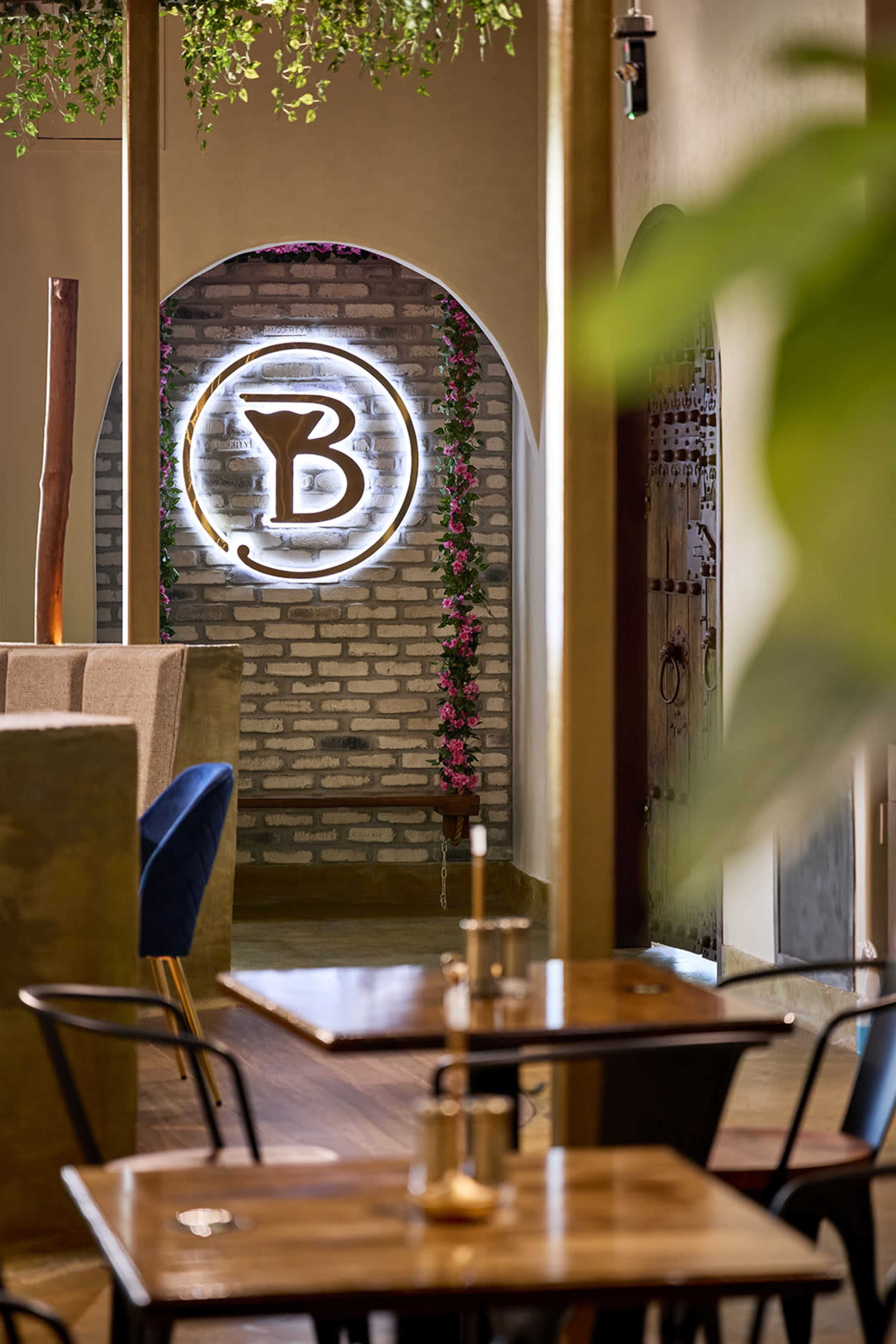 The image shows a modern restaurant interior featuring a brick wall with a circular neon sign, surrounded by greenery and wooden furnishings.