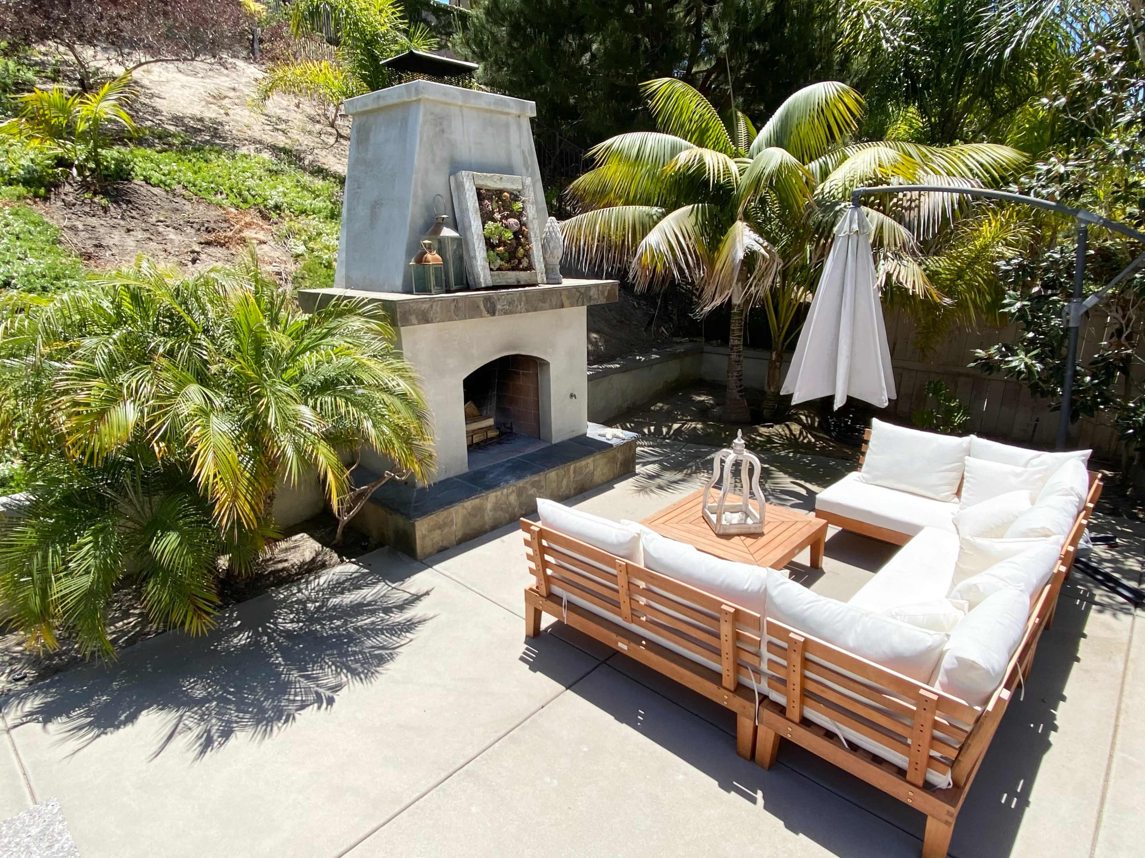 A patio area with a sectional sofa, a coffee table, an outdoor fireplace, and a large umbrella next to tropical plants and trees.