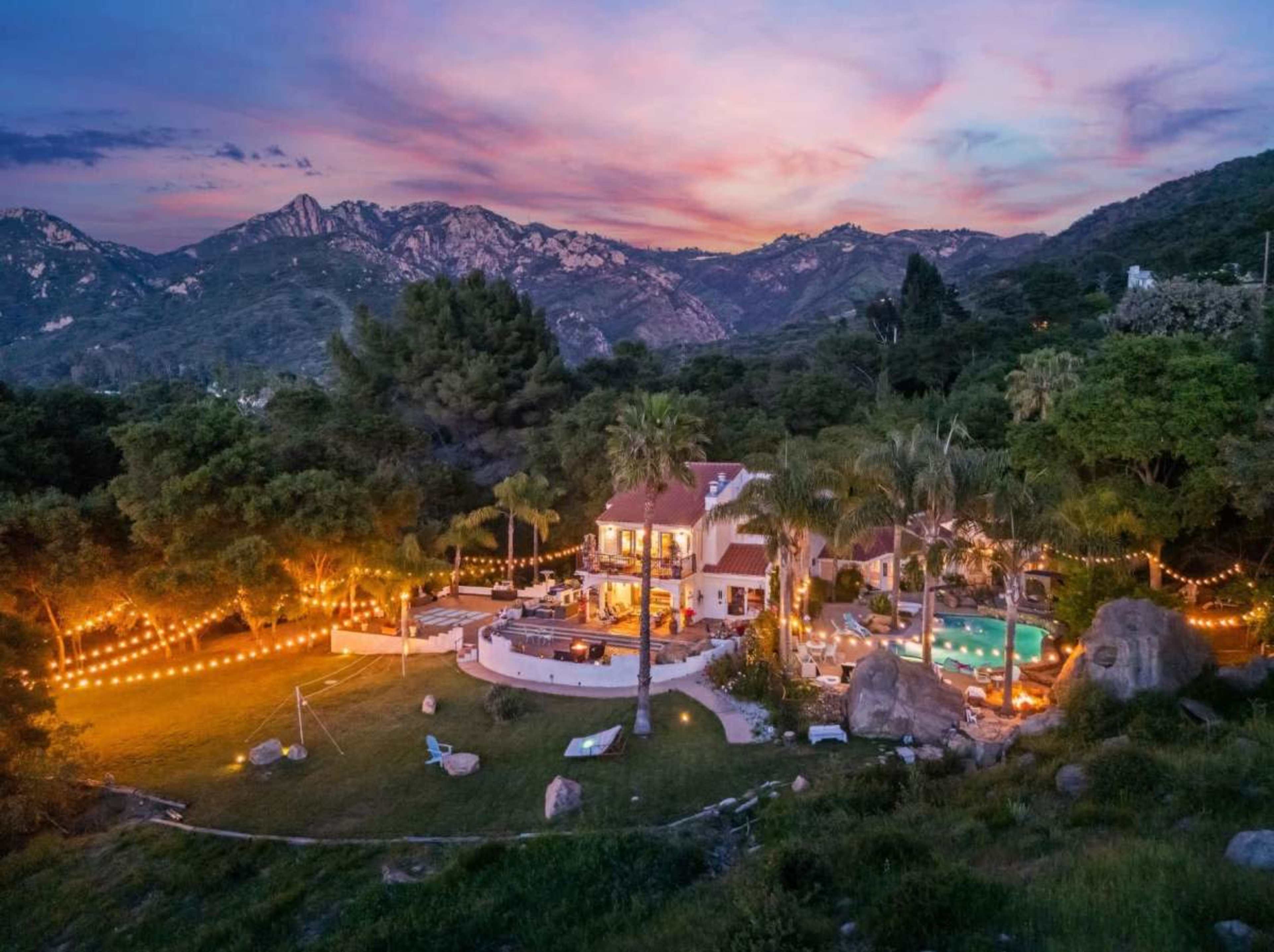 The image depicts a spacious house surrounded by mountains, with a pool and string lights illuminating the outdoor area during twilight.