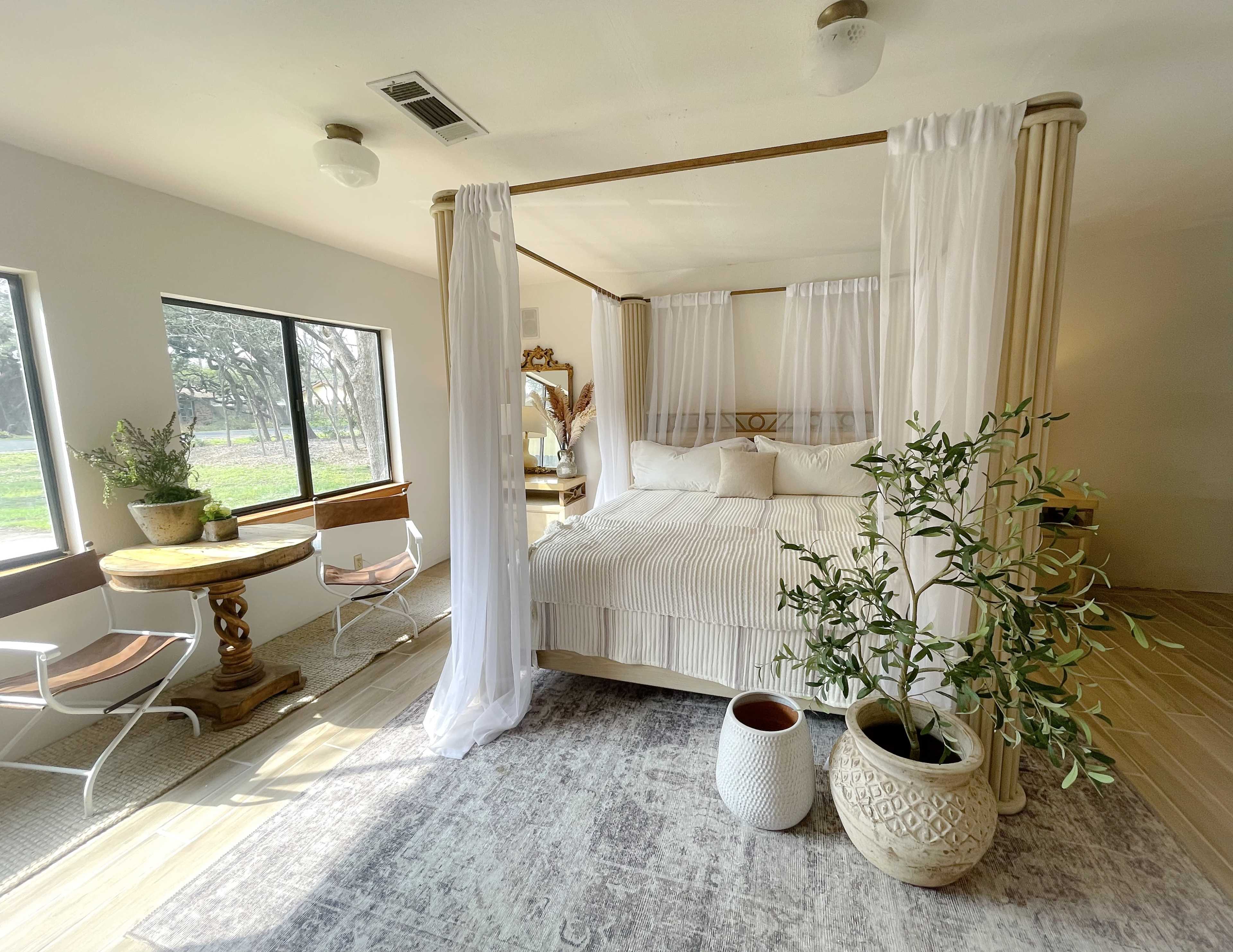A bright, airy bedroom features a four-poster bed with sheer drapes, a small circular dining table, and potted plants.