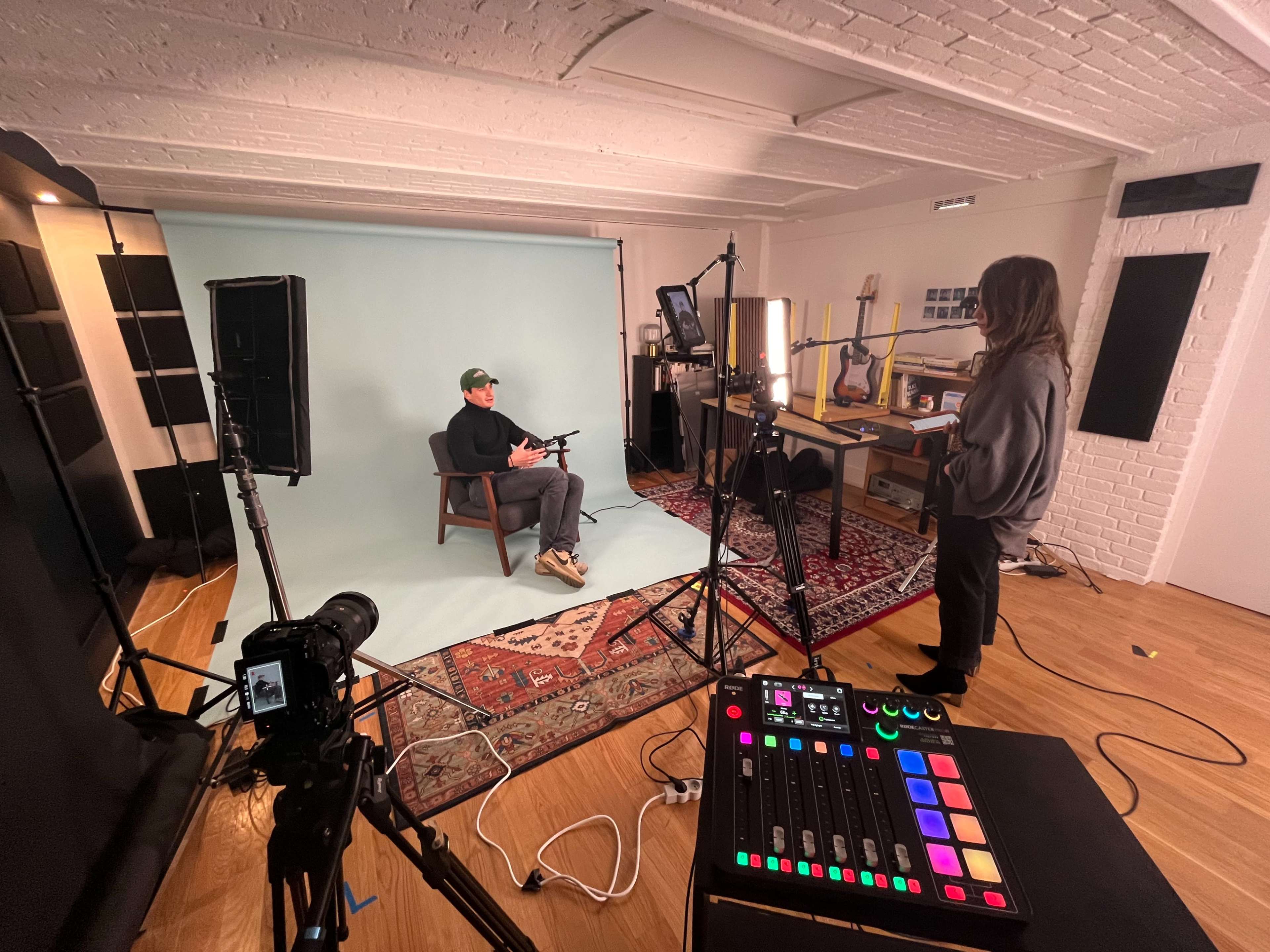 A man sits on a chair in front of a light blue backdrop while a woman stands nearby, surrounded by filming equipment and studio lights.