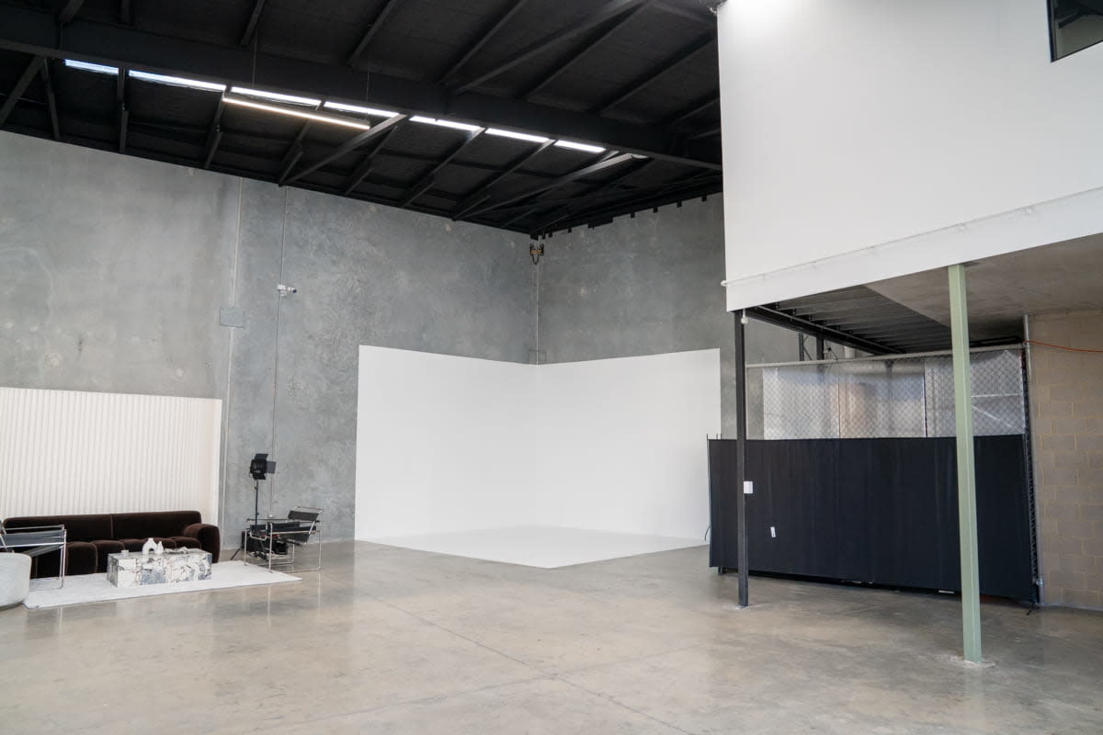 The image shows a spacious, unfinished industrial room with a large white backdrop on one side, a dark partition, and a seating area featuring a sofa and table.