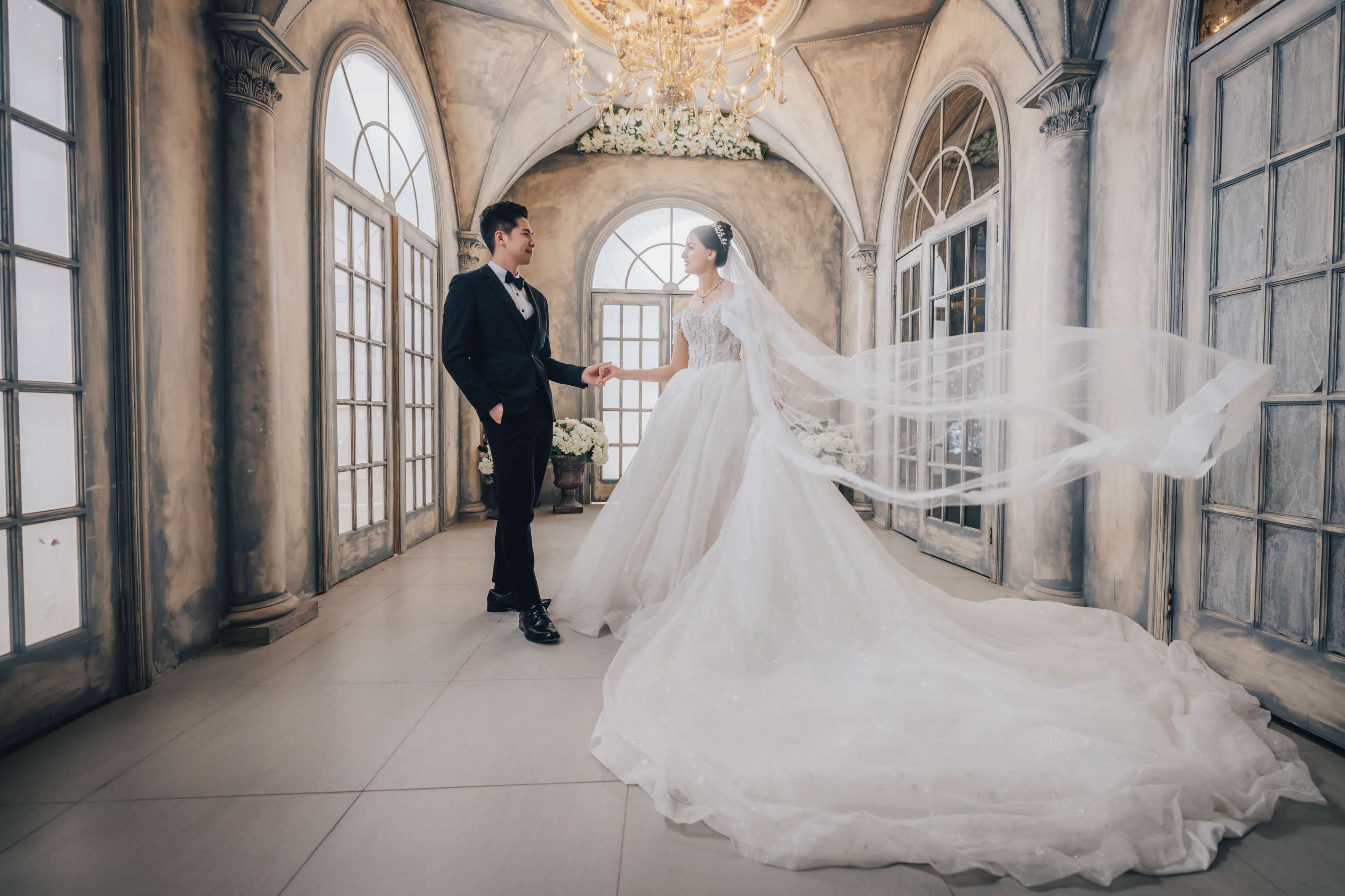 A bride in a flowing gown and a groom in formal attire stand together in an elegantly decorated indoor space with large windows and a chandelier.