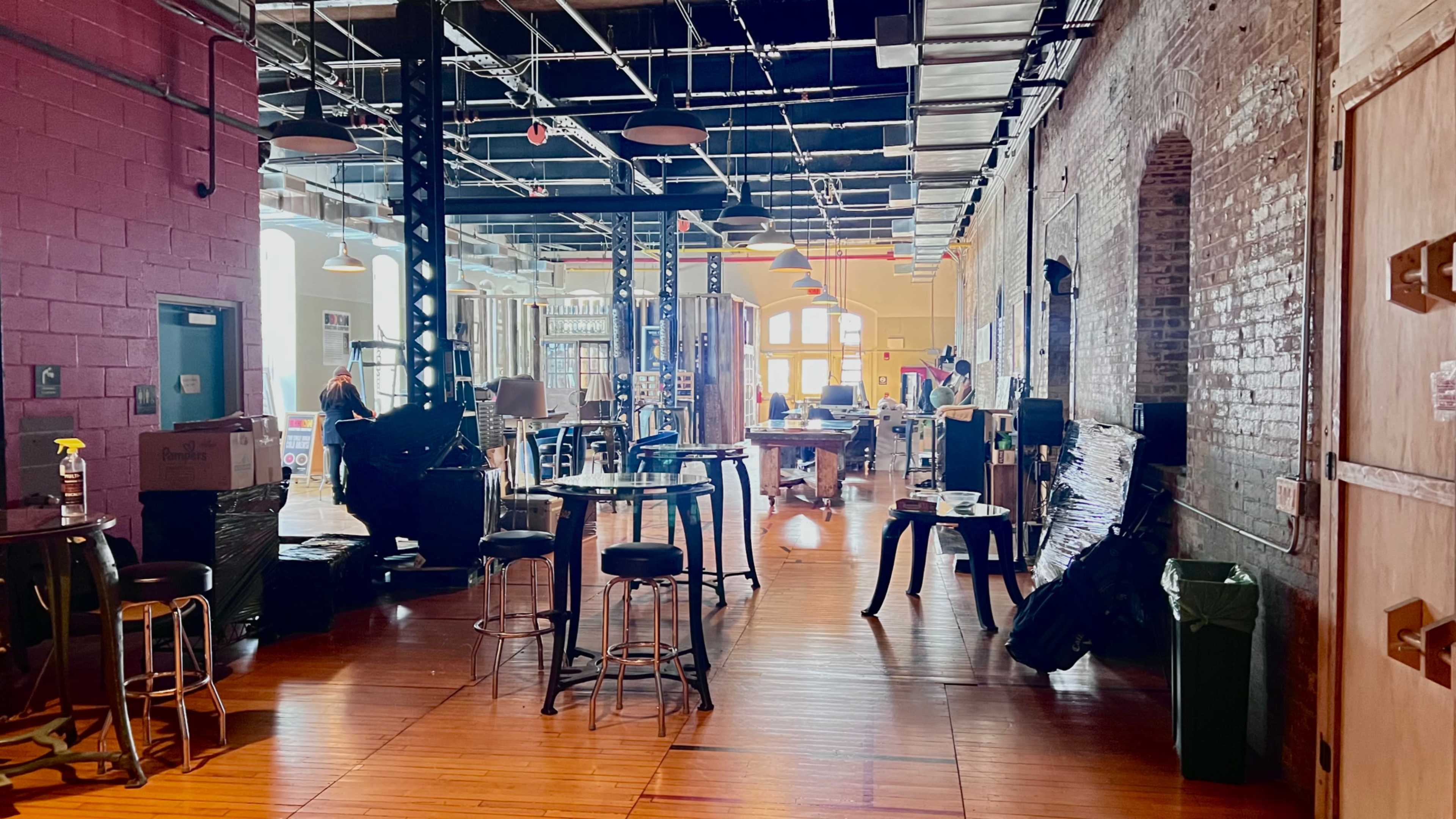 The image shows an open space with wooden floors, tall ceilings, and various tables and chairs, surrounded by boxes and workstations.