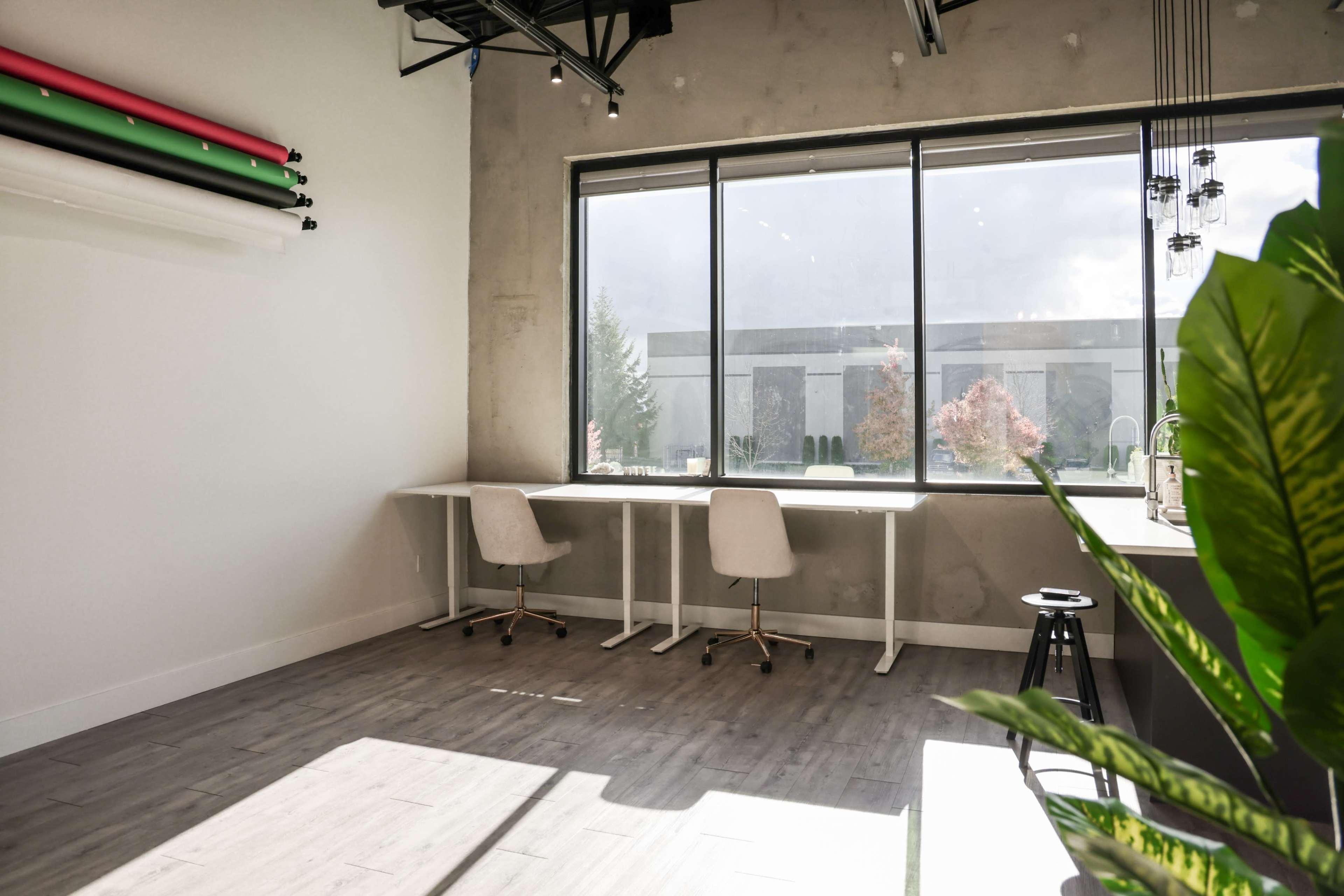 A well-lit workspace features two desks with chairs beside a large window overlooking an outdoor area.