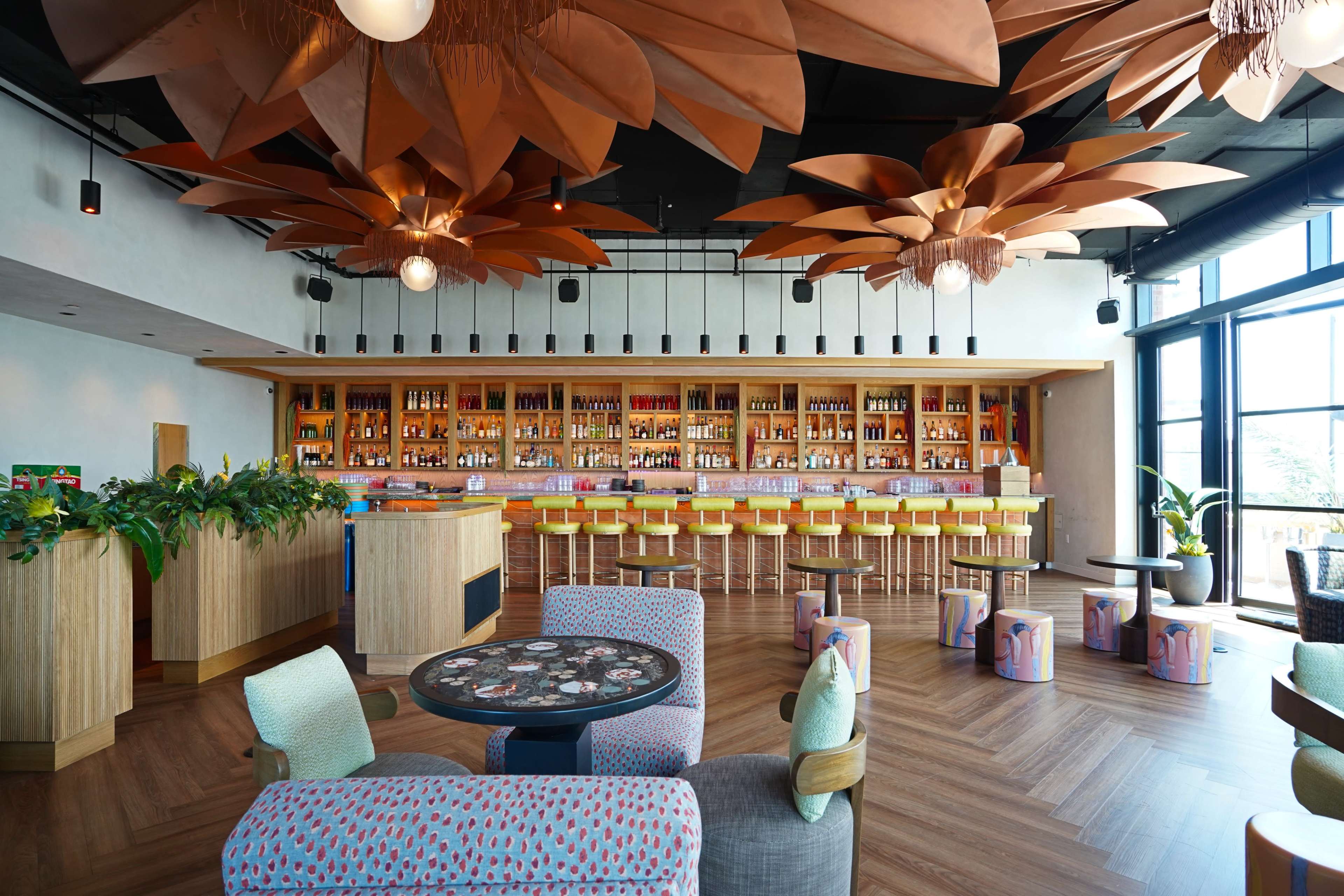 The image features a modern bar interior with large floral light fixtures, a wooden bar counter lined with colorful stools, and vibrant plants positioned throughout the space.