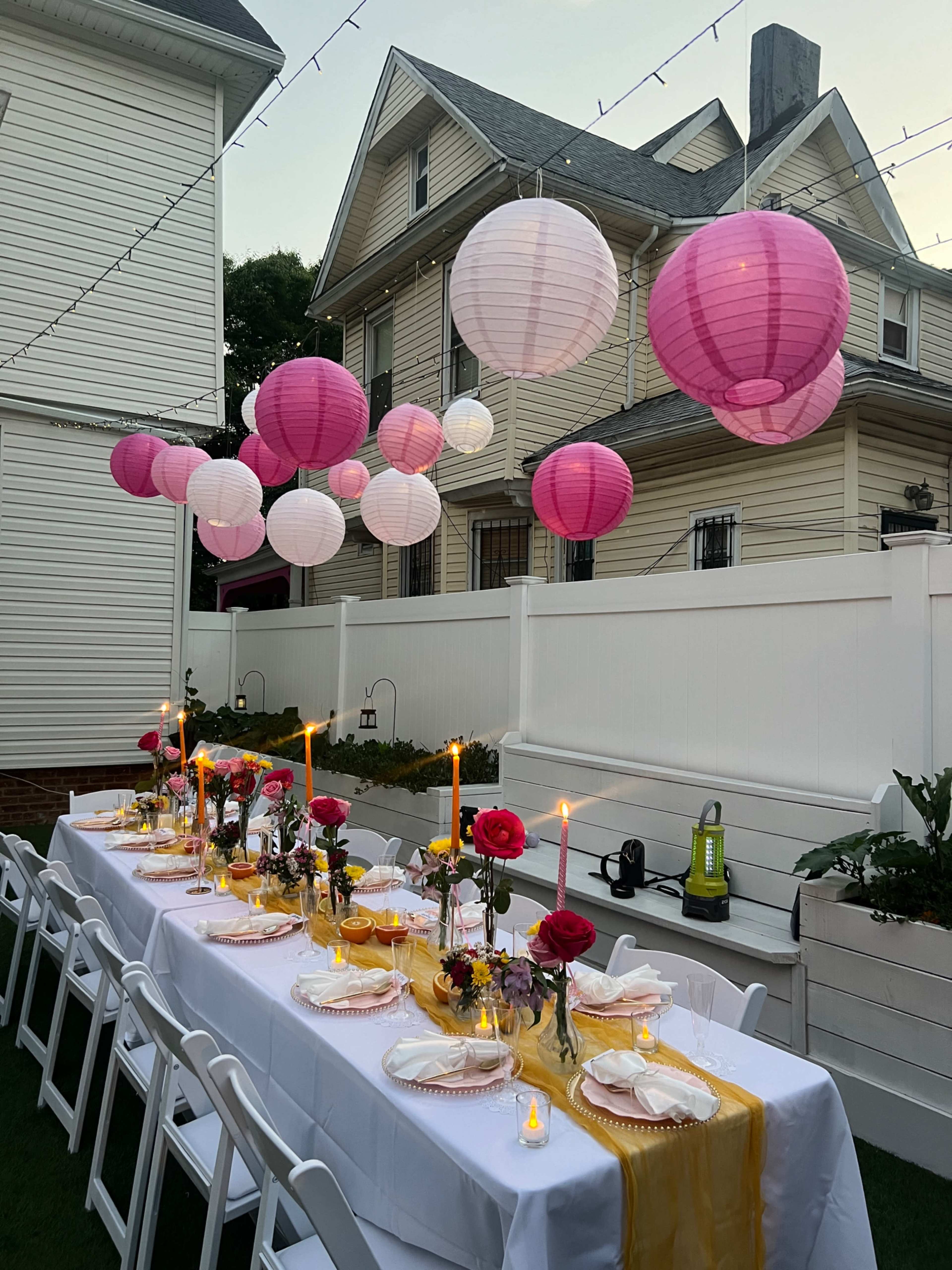 A long dining table adorned with candles and flowers is set outdoors under decorative pink lanterns, against the backdrop of adjacent houses and a white fence.