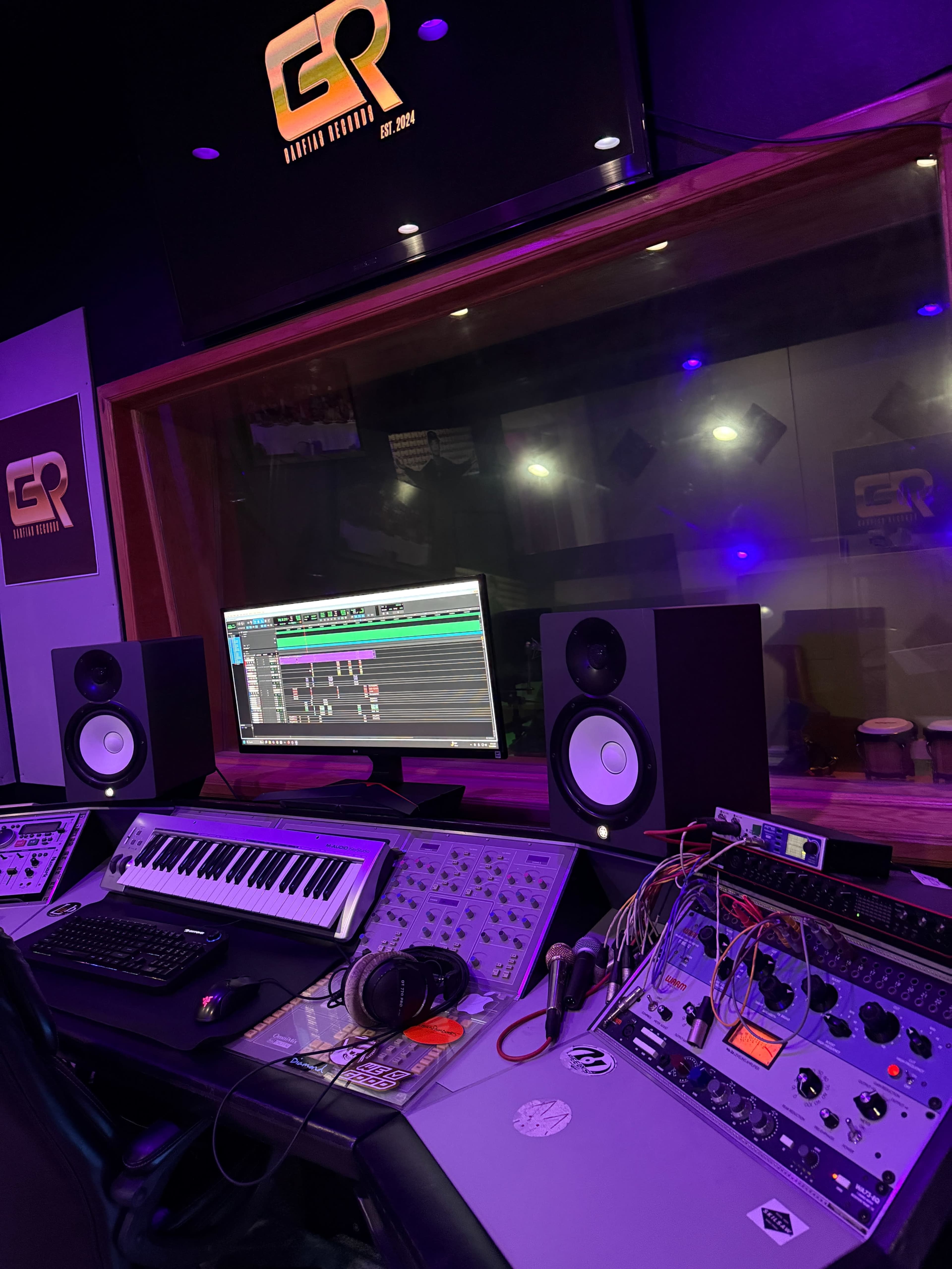 The image shows a music studio with a computer monitor displaying audio software, a keyboard, studio monitors, and various audio equipment arranged on a control desk.