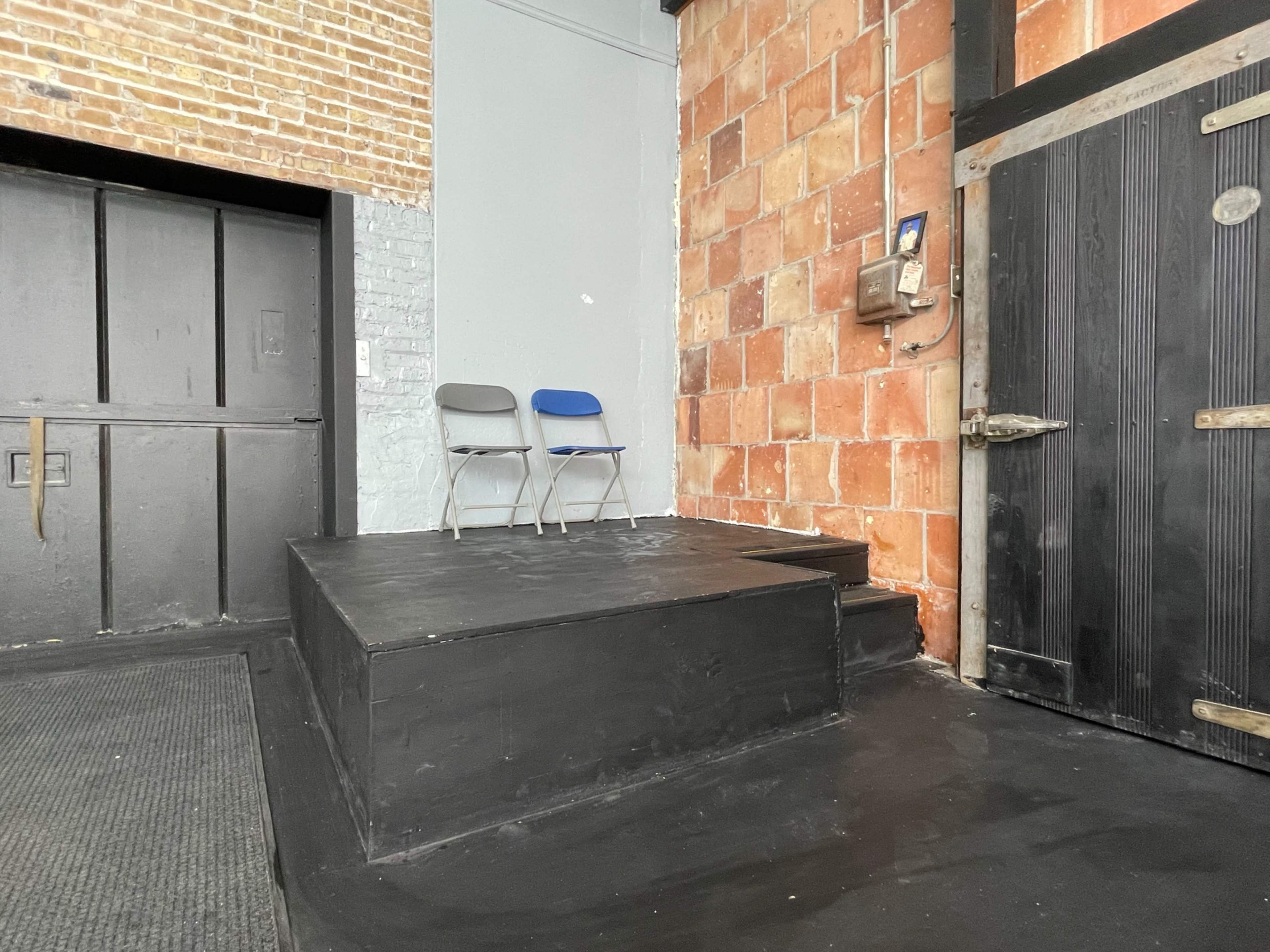 The image shows a small, elevated platform with two blue chairs positioned on it, adjacent to a brick wall and a large black door.