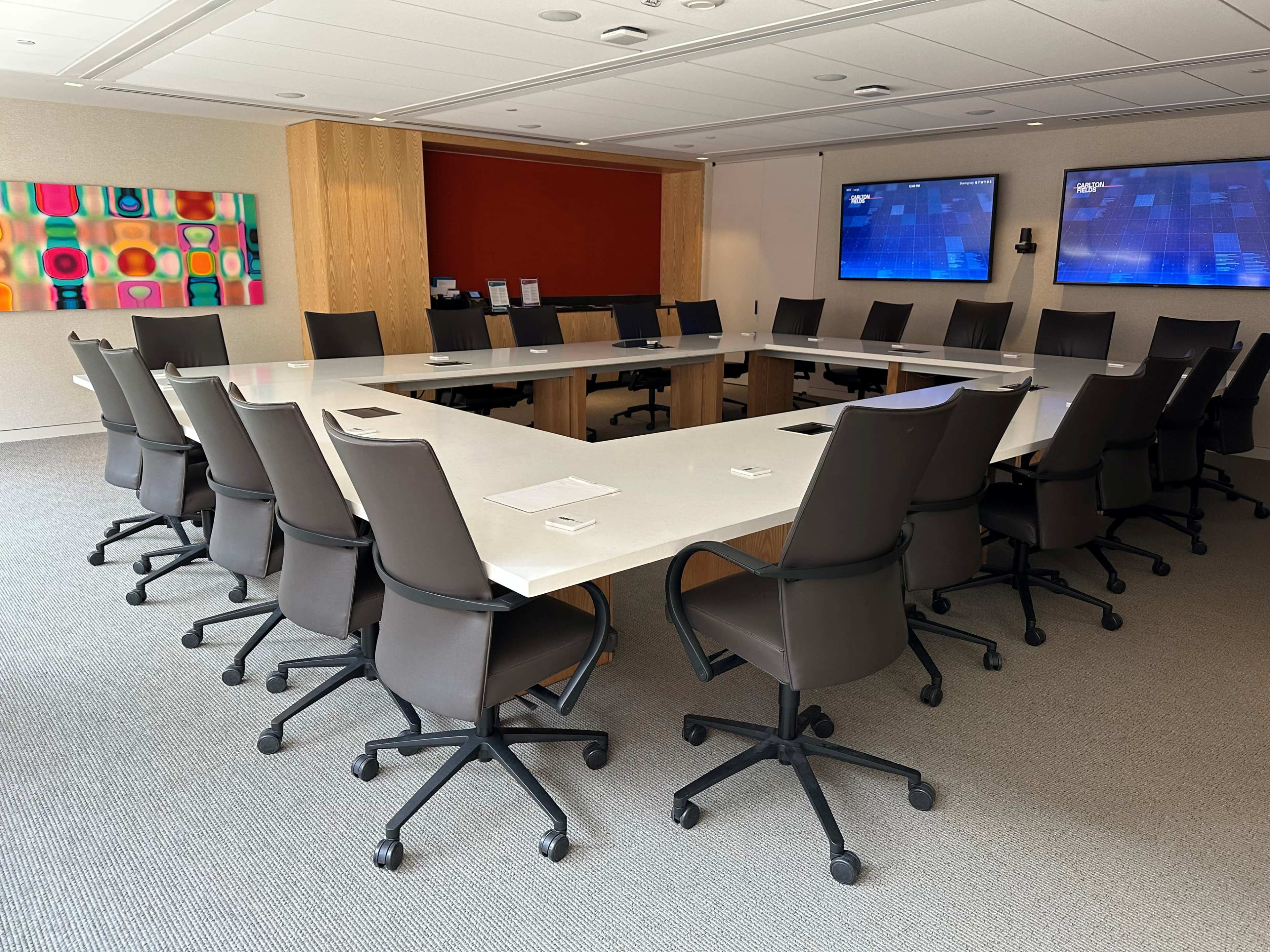 The image shows a large conference room with a rectangular table surrounded by ergonomic office chairs, equipped with two screens on the walls and a colorful abstract artwork.