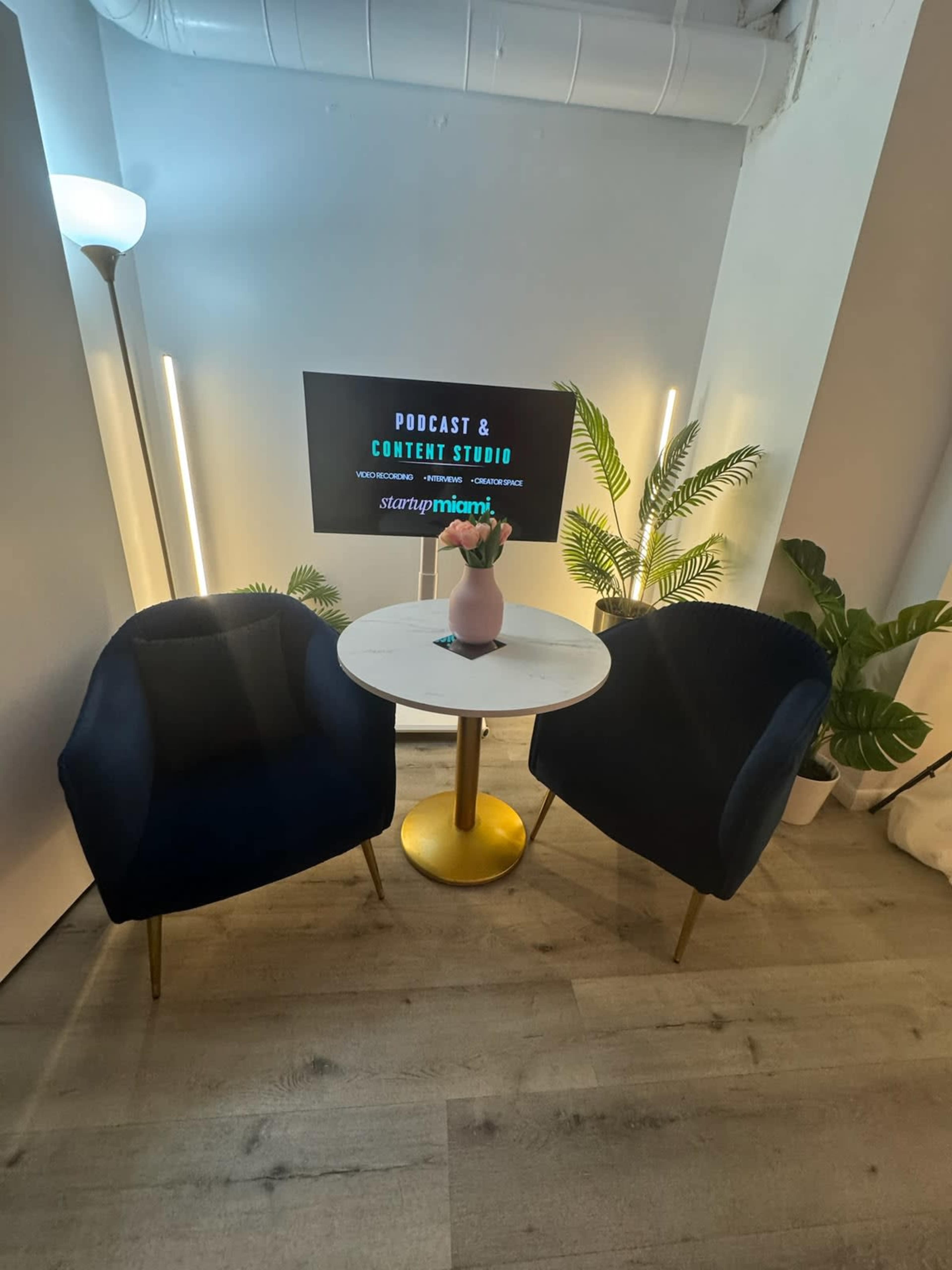 The image shows a podcast and content studio setup with two navy blue chairs, a small round table, and a screen displaying "Podcast & Content Studio" in a well-lit, modern environment.