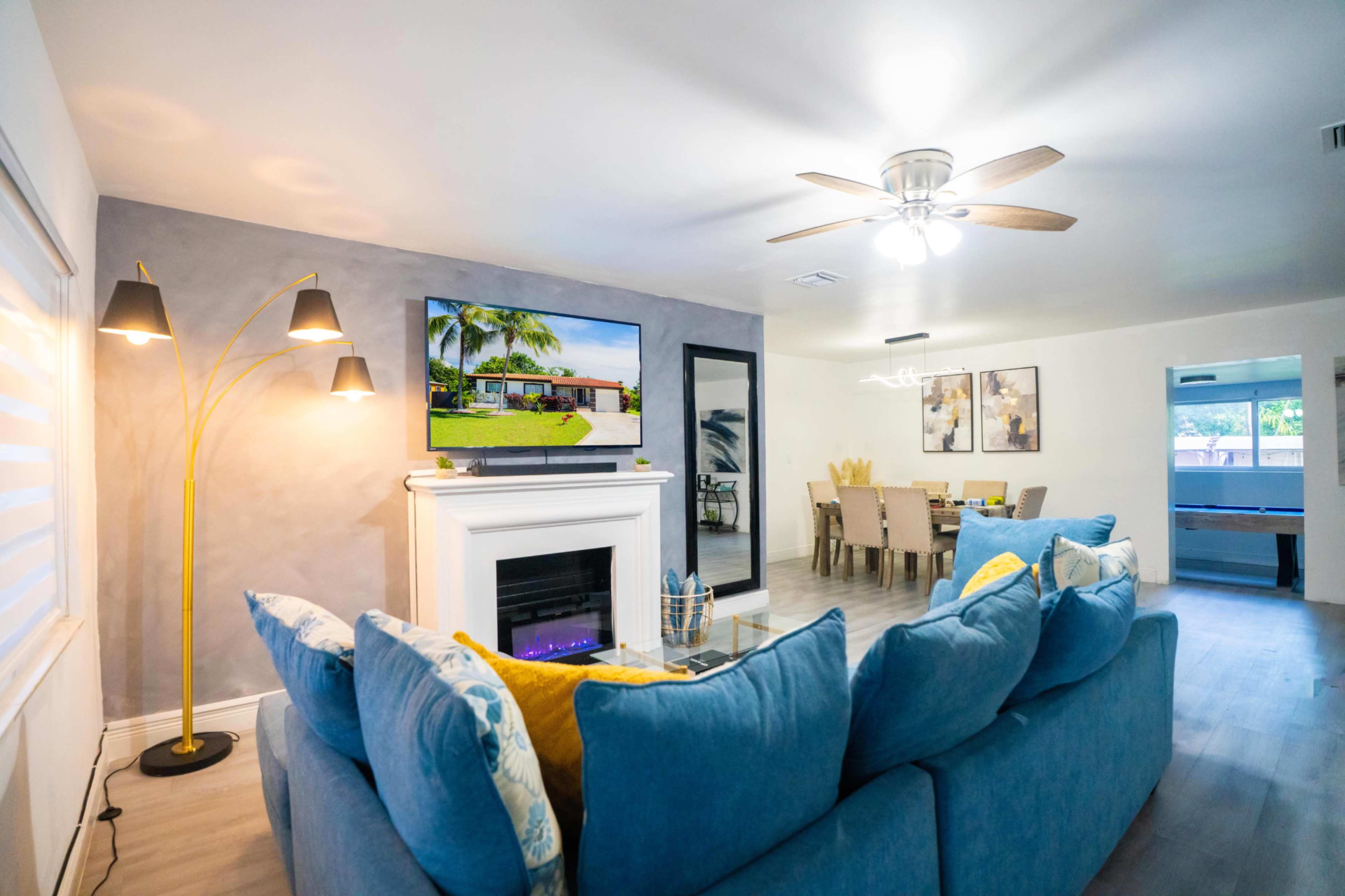 The living room features a blue sectional couch facing a mounted television above a fireplace, with a dining area visible in the background.