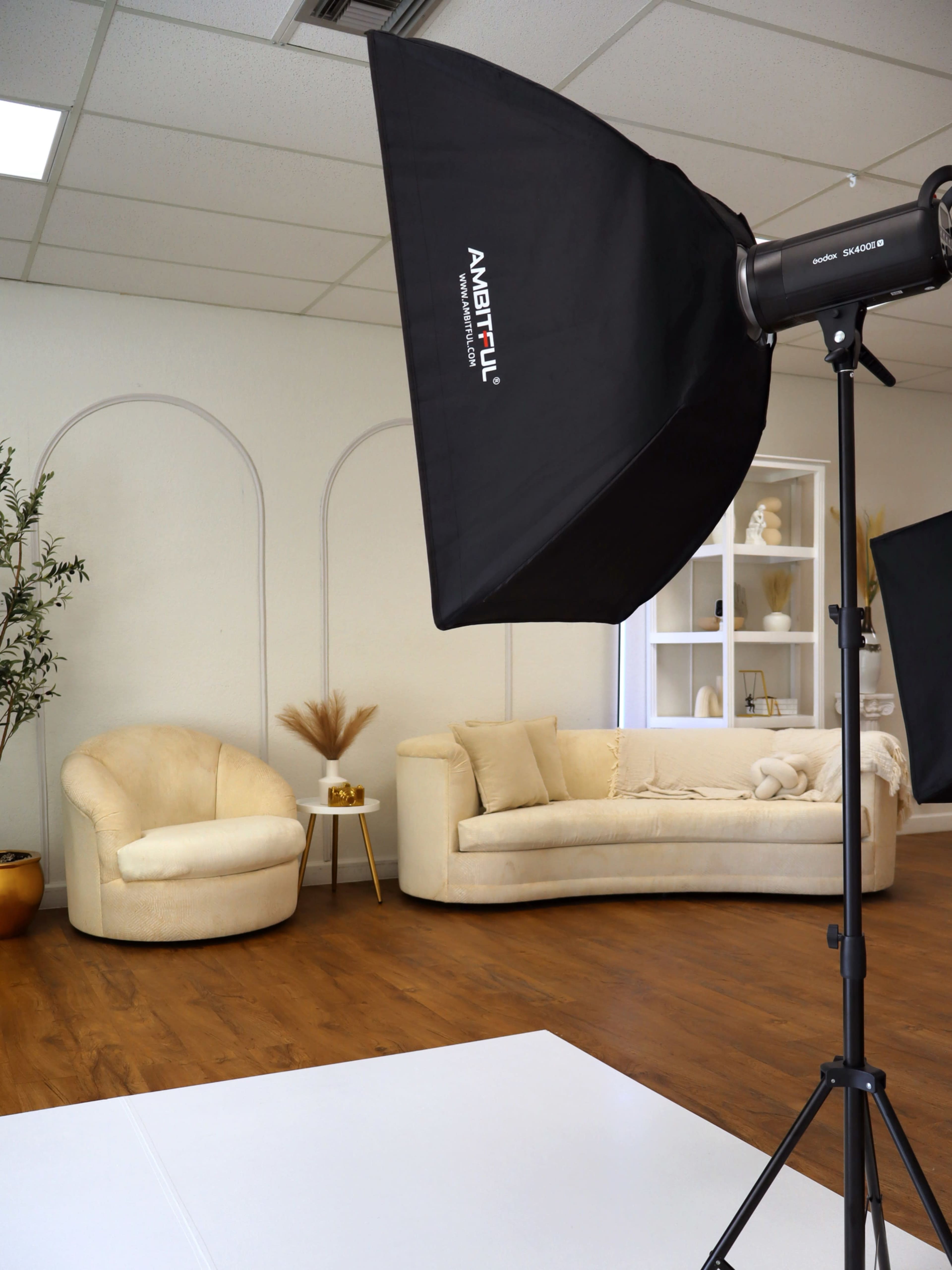 The image shows a well-lit photography studio featuring a cream-colored sofa, a cozy armchair, and light-colored decor elements.