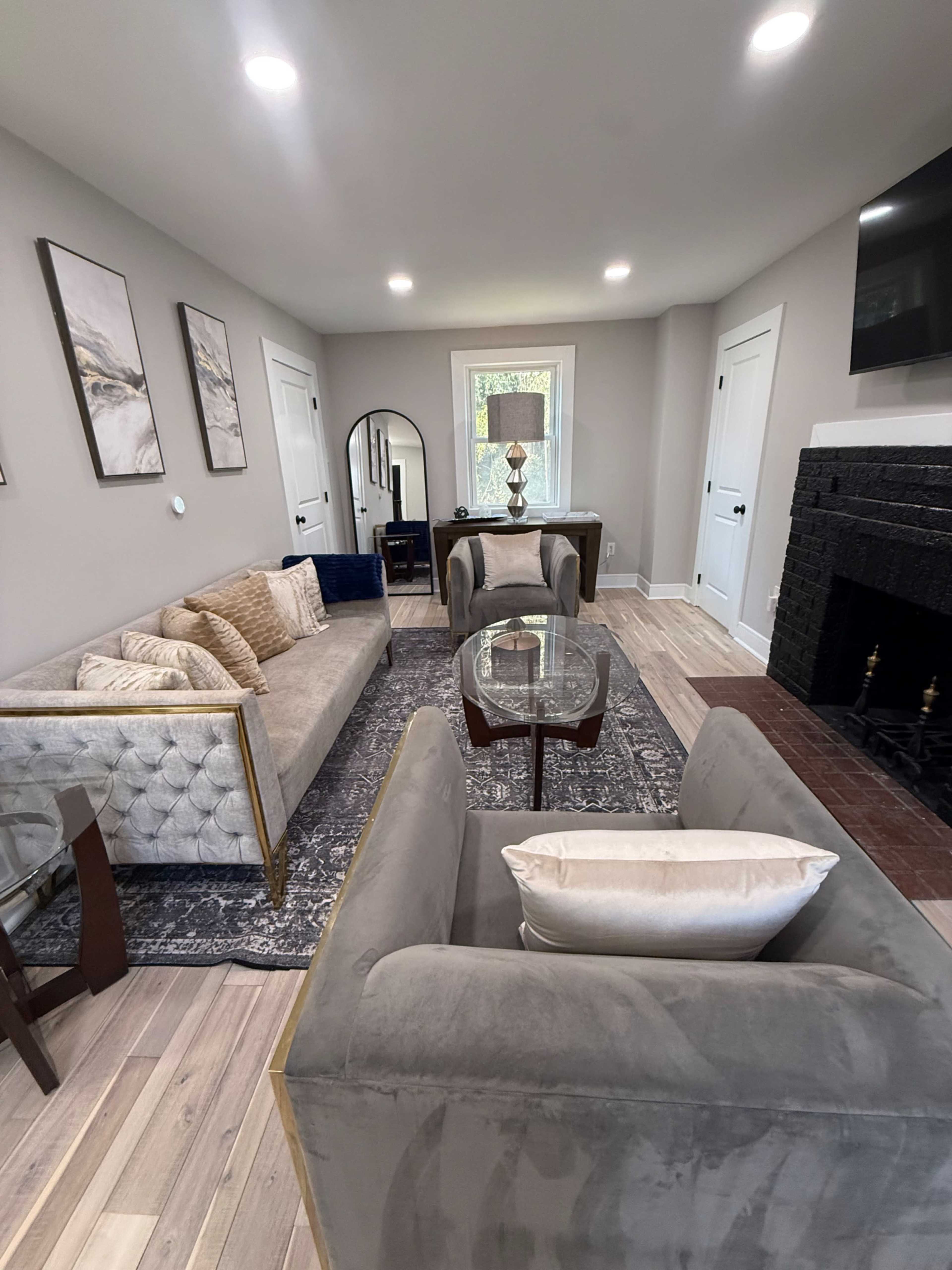 The image shows a modern living room with two upholstered chairs, a sofa, a glass coffee table on a patterned rug, and a fireplace, all illuminated by bright ceiling lights.