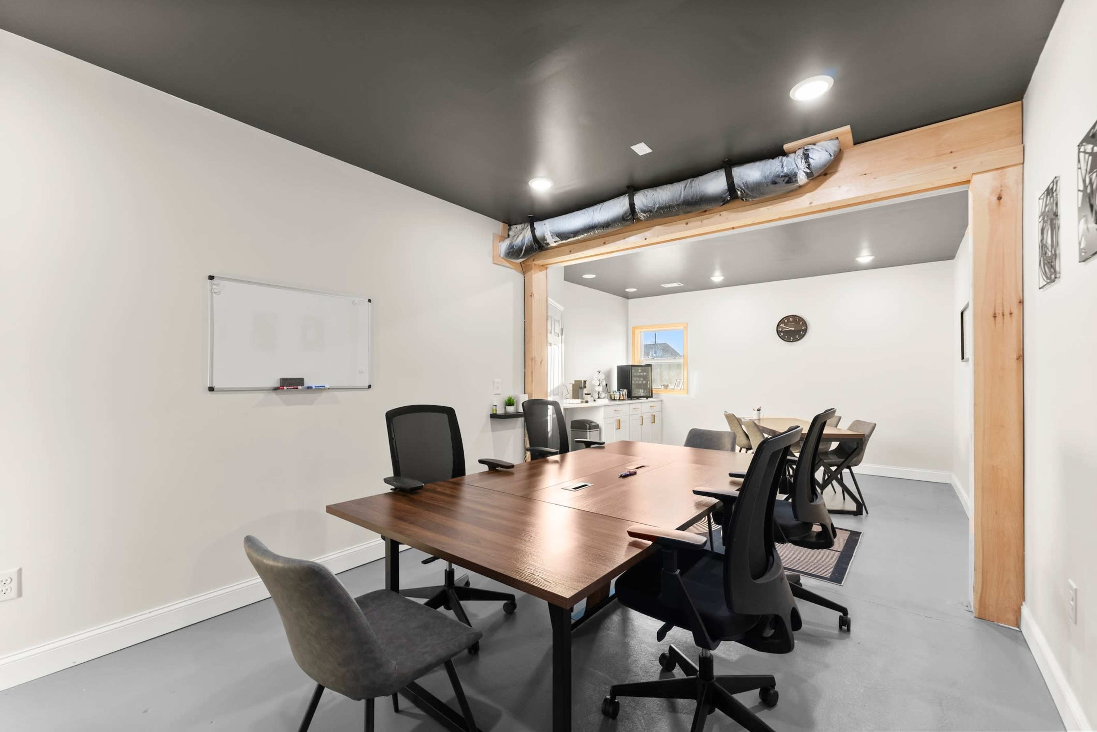 The image shows a modern conference room featuring a large wooden table surrounded by several chairs, with a kitchenette area and a whiteboard on the wall.