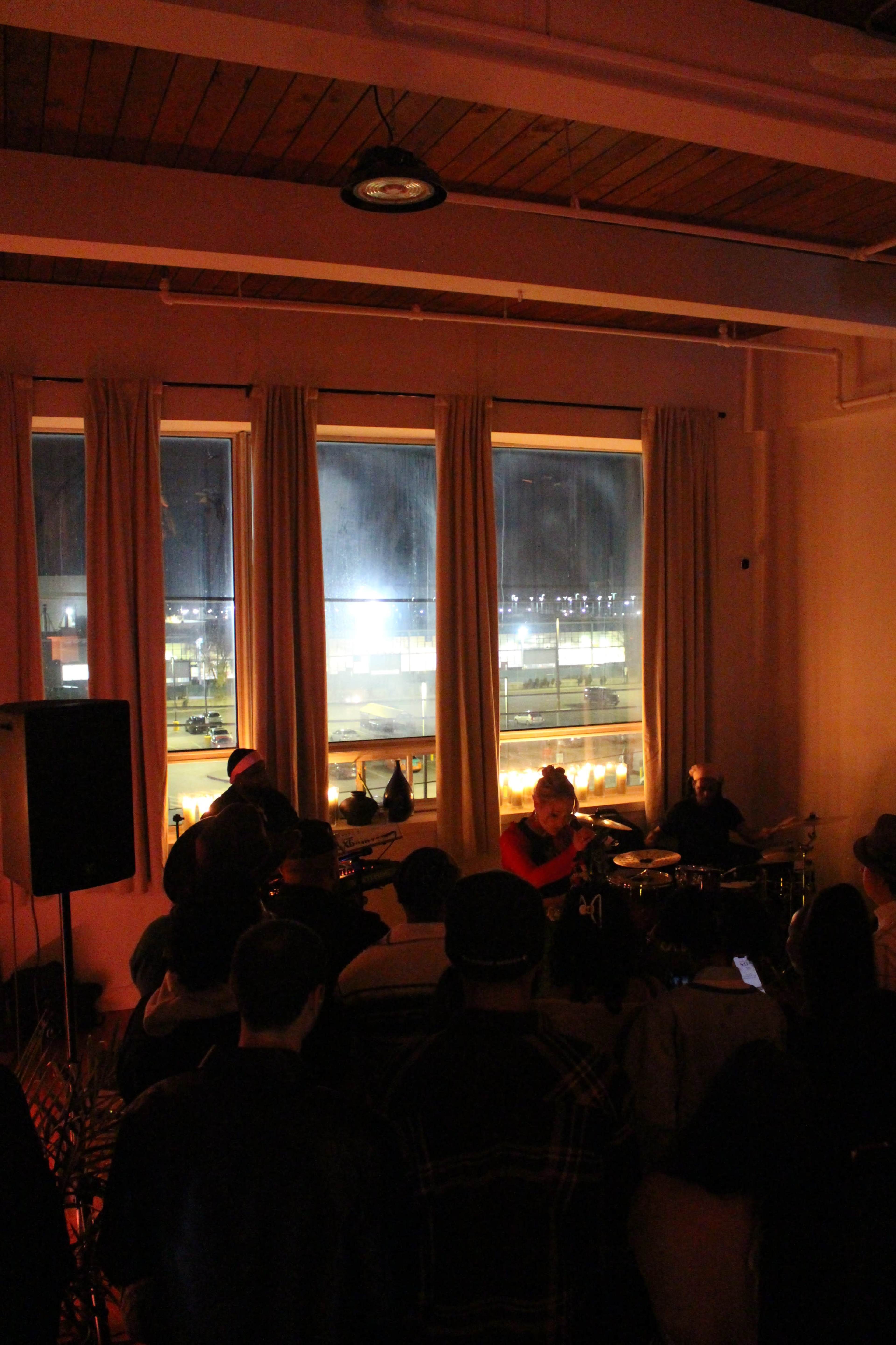 A small crowd watches a live music performance indoors, with large windows revealing a brightly lit outdoor scene.