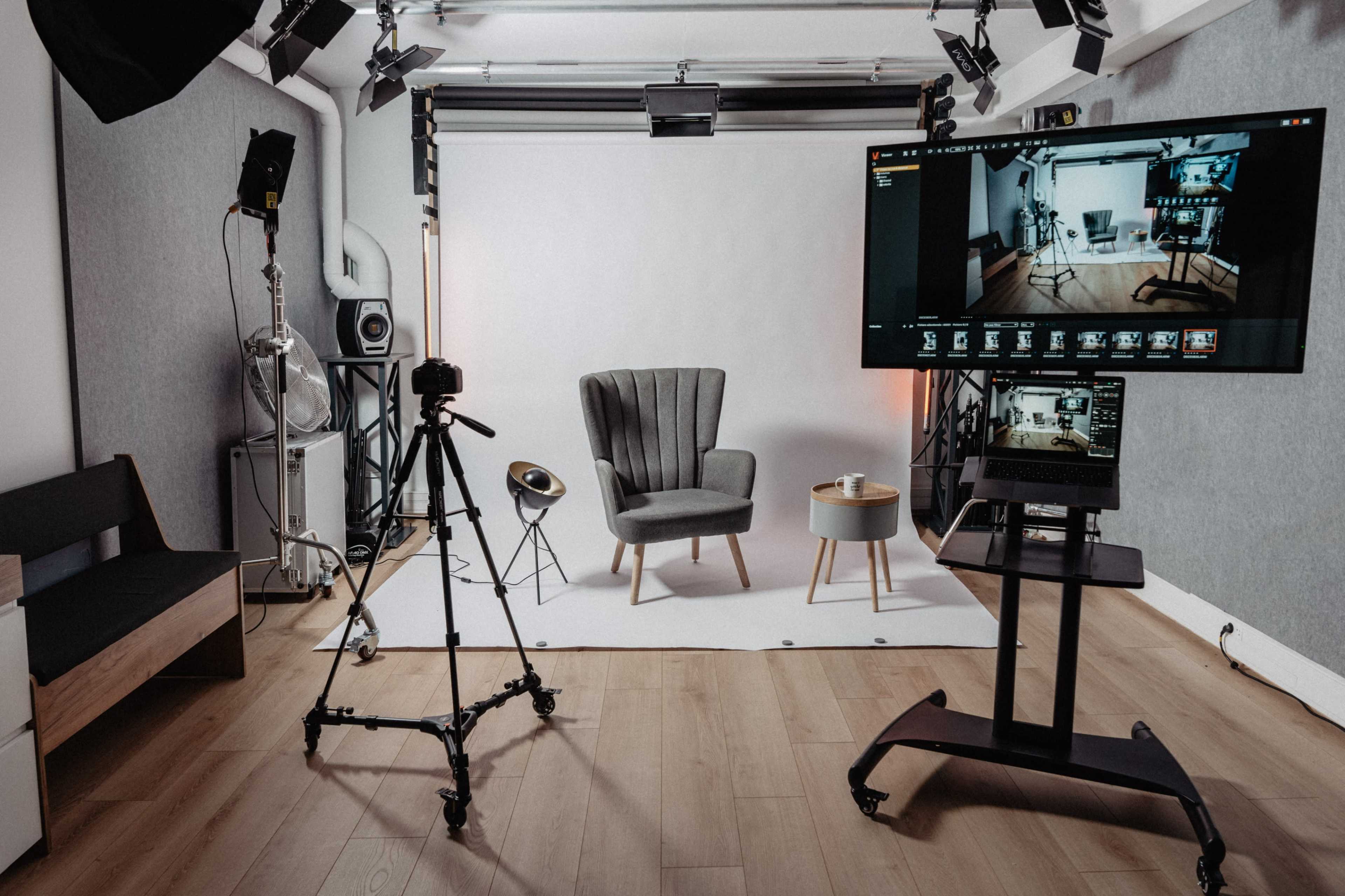 A photography studio equipped with a tripod, lighting setup, a gray armchair, a small table, and two screens displaying camera settings.