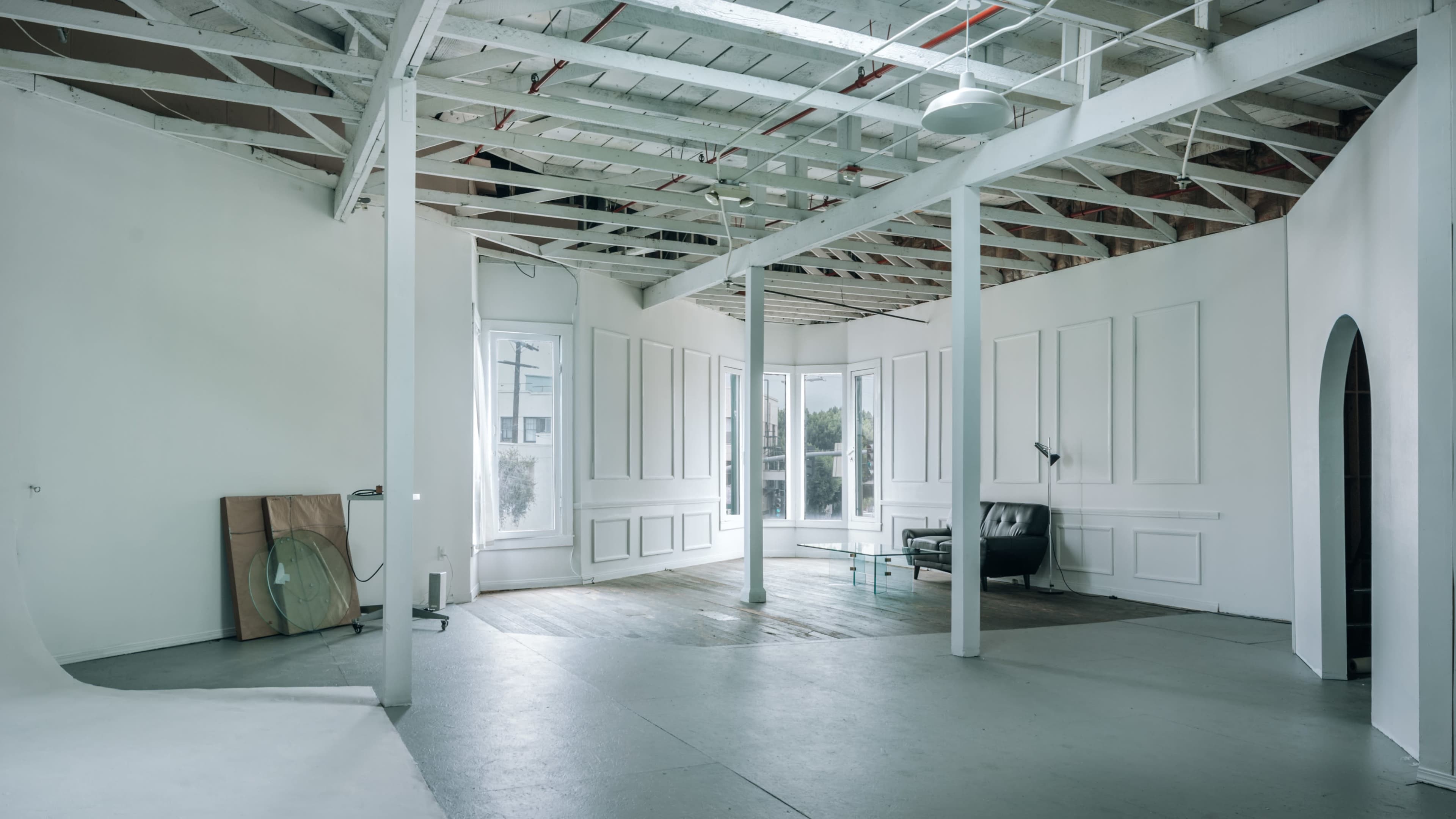 A spacious, empty room with white walls, a wooden floor, exposed beams, and large windows on one side.