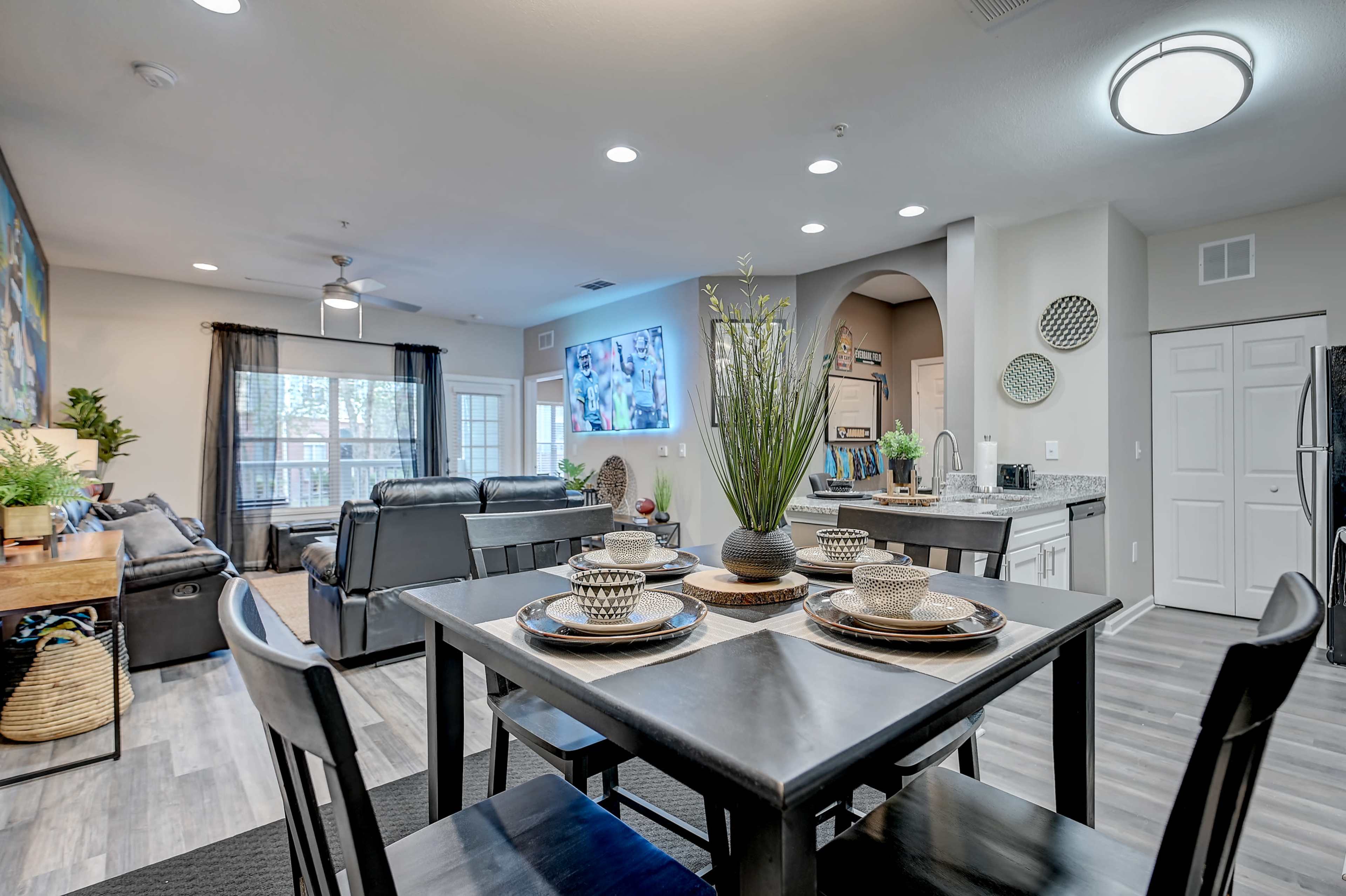 The image shows a spacious, modern living area featuring a dining table set for four, a seating area with a sectional sofa, and a wall-mounted television.