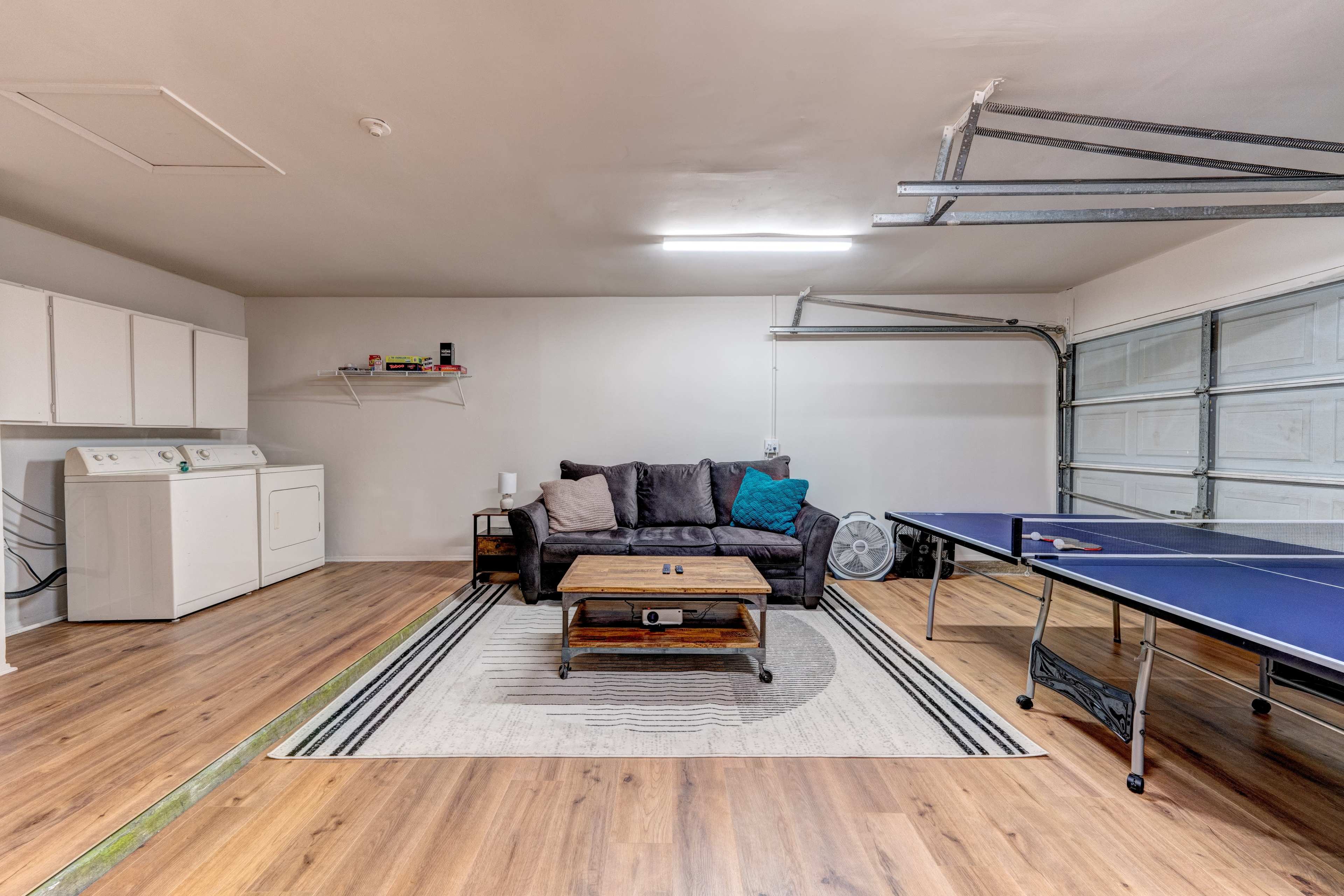 A garage features a couch and coffee table on a rug, a laundry area with a washer, and a ping pong table beside a garage door.
