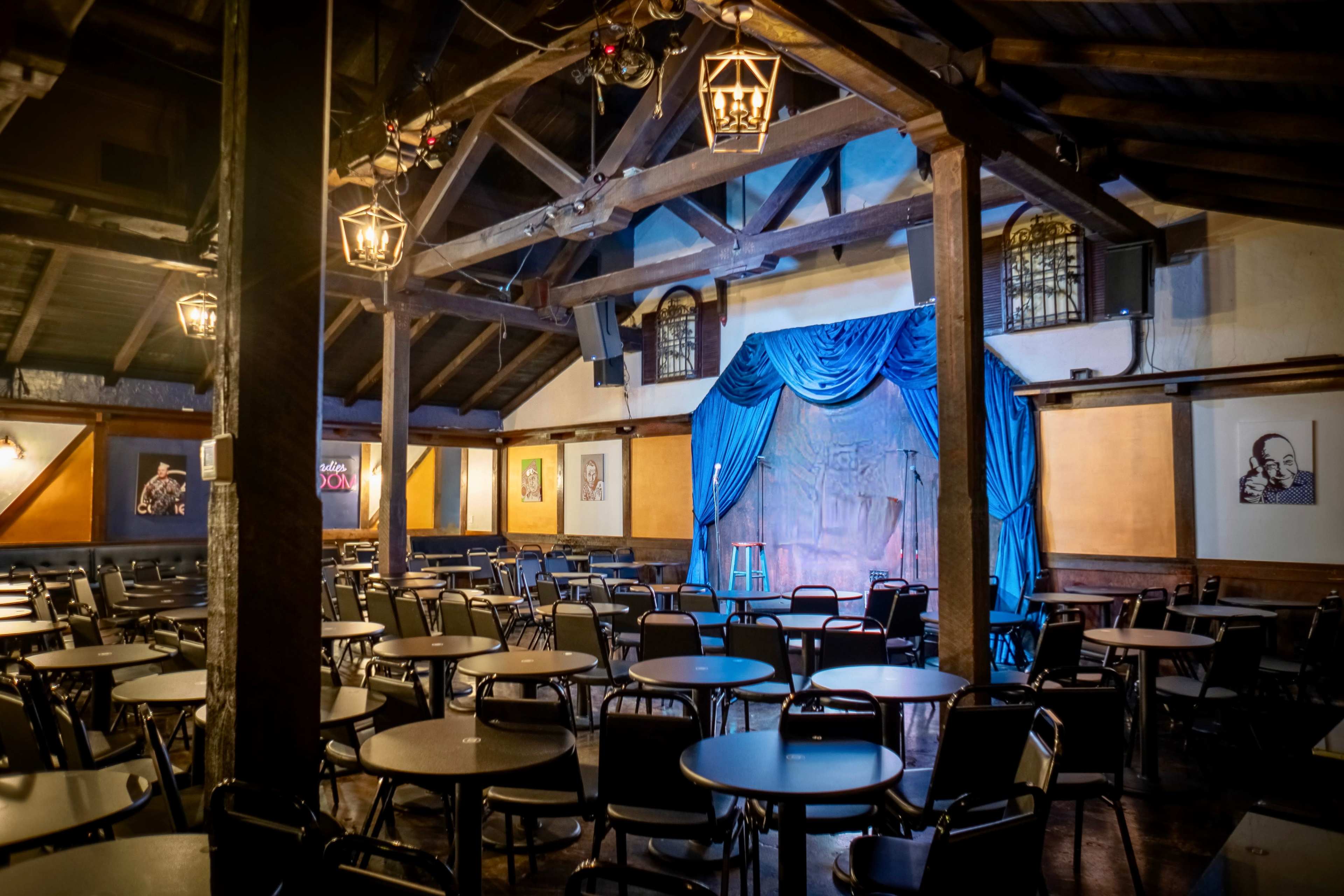 A dimly lit comedy club features a stage with blue drapery and empty tables arranged in rows around the room.