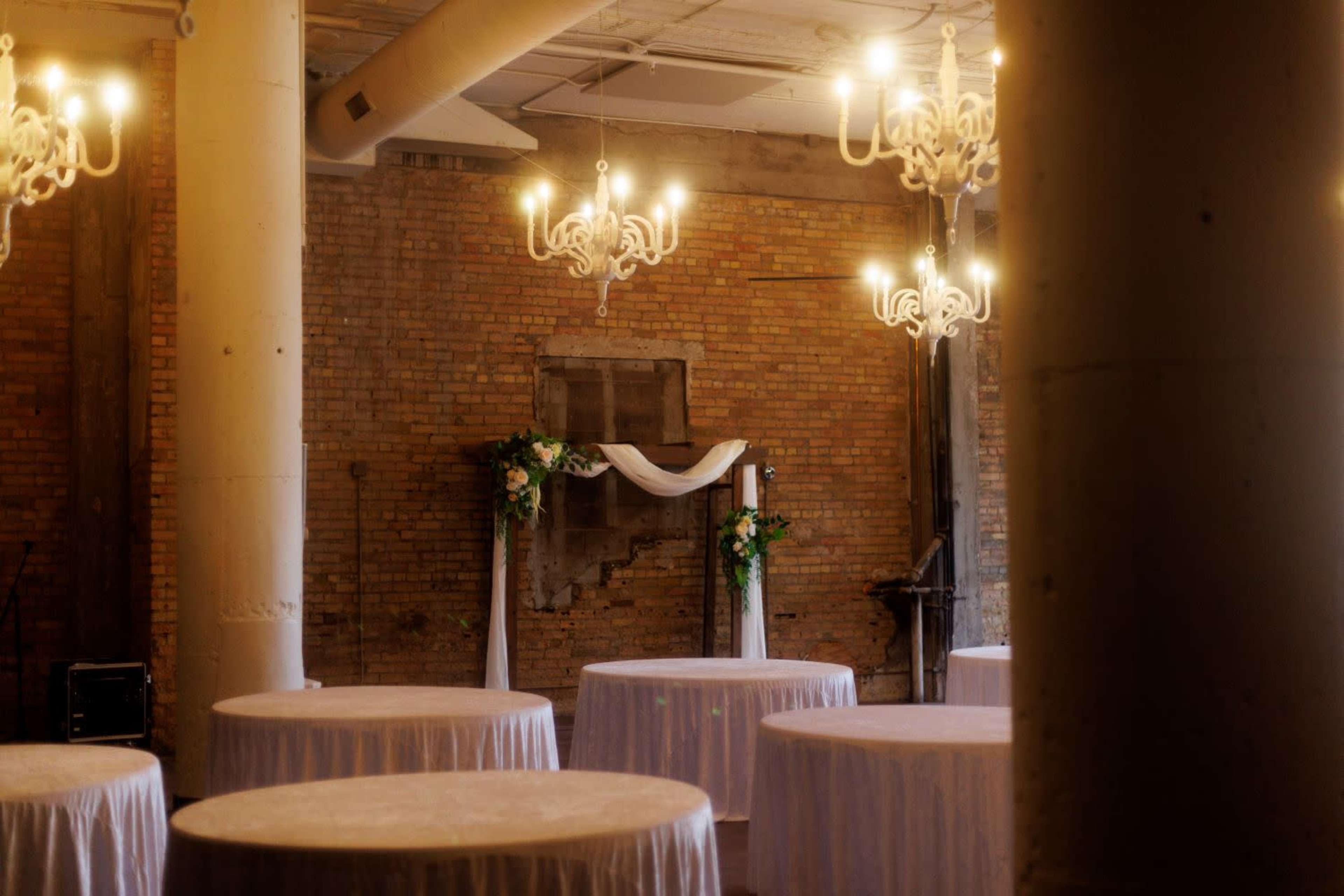 The image shows a decorated indoor venue featuring several round tables with white tablecloths and chandeliers hanging from the ceiling.