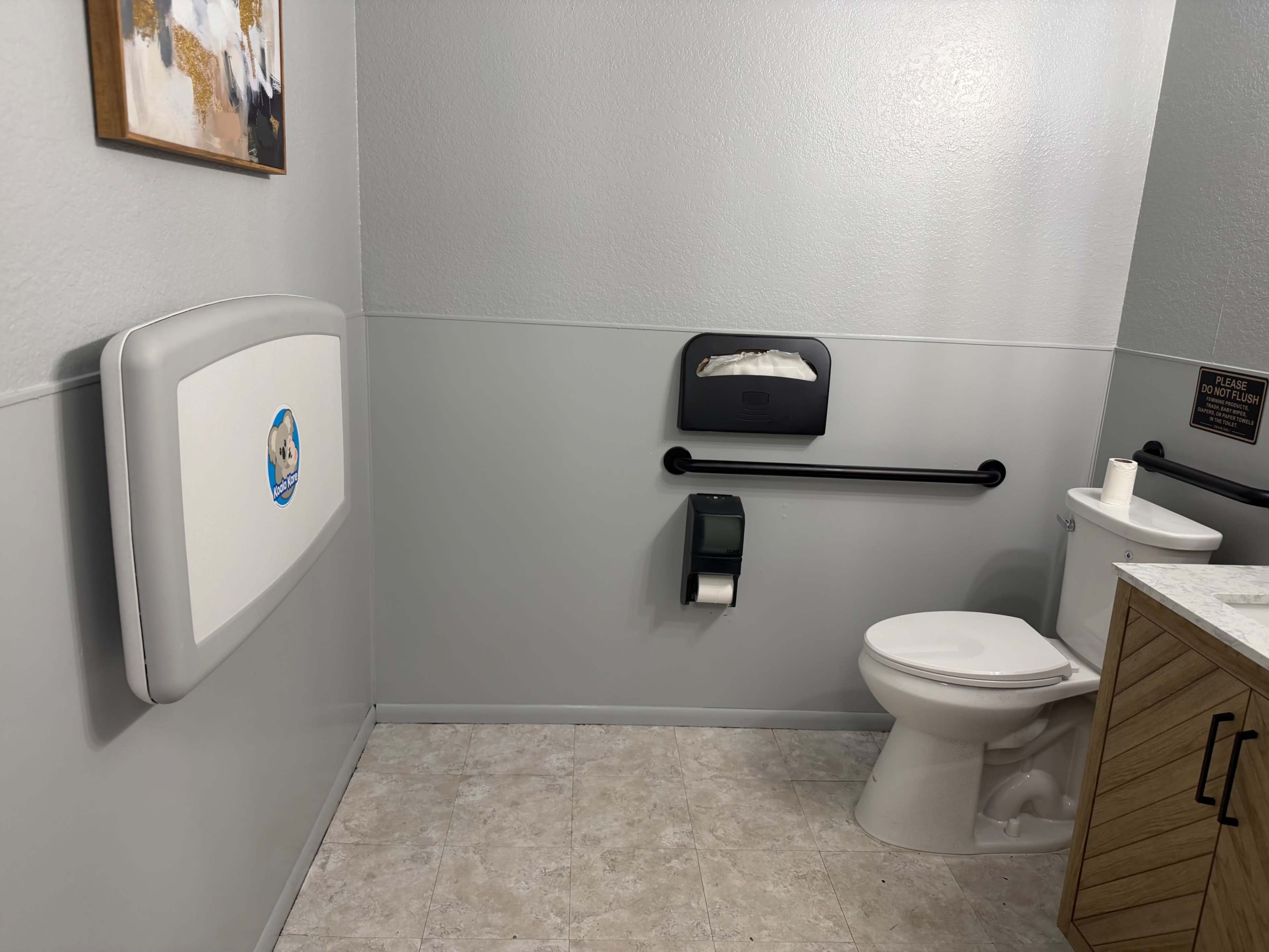 The image shows a clean restroom featuring a toilet, a wall-mounted baby changing station, and a wall-mounted paper towel dispenser on gray walls with tile flooring.