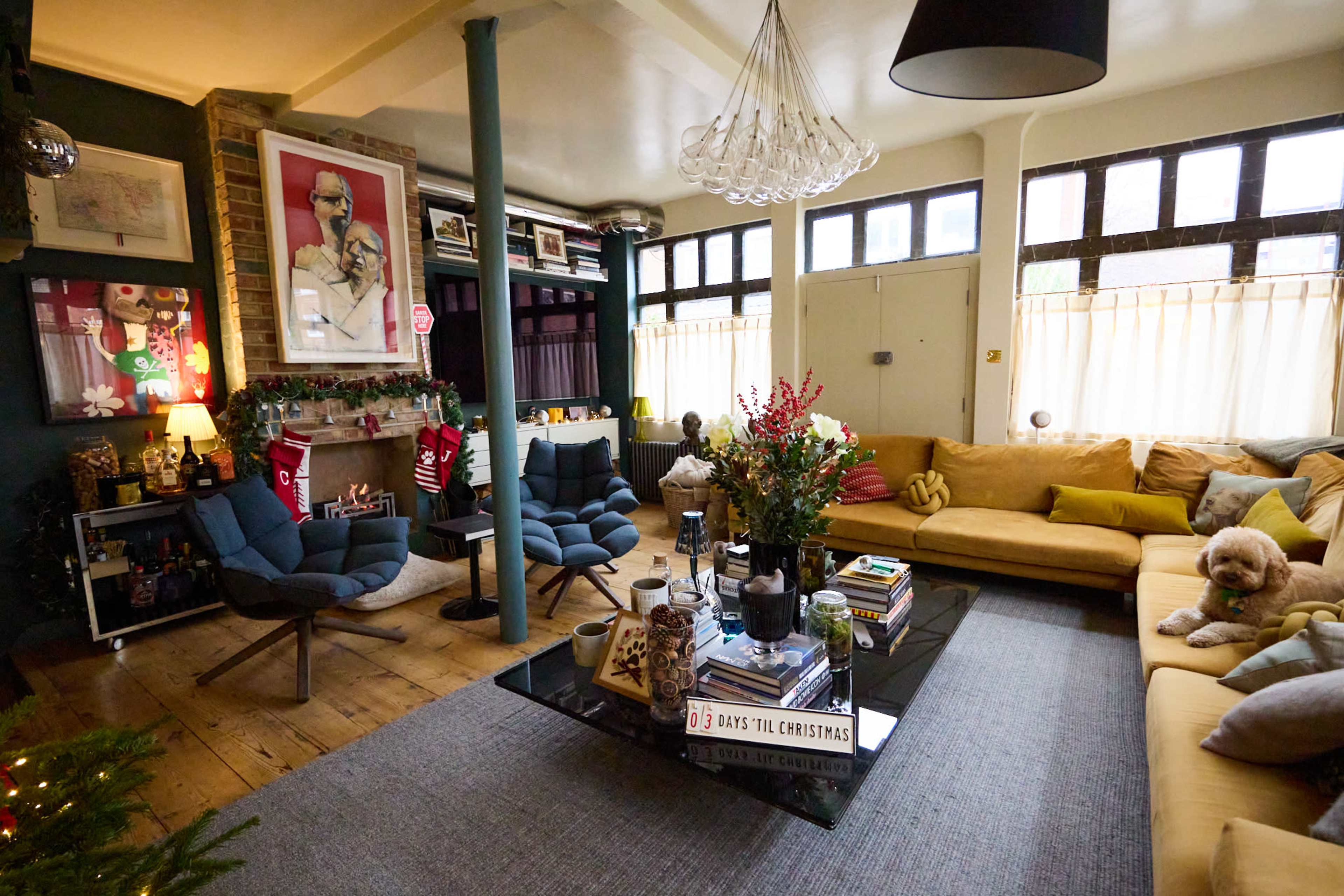 A cozy living room features a mix of modern and vintage furniture, including two lounge chairs and a large yellow sofa, with a coffee table adorned with books and decor, and a Christmas countdown display.