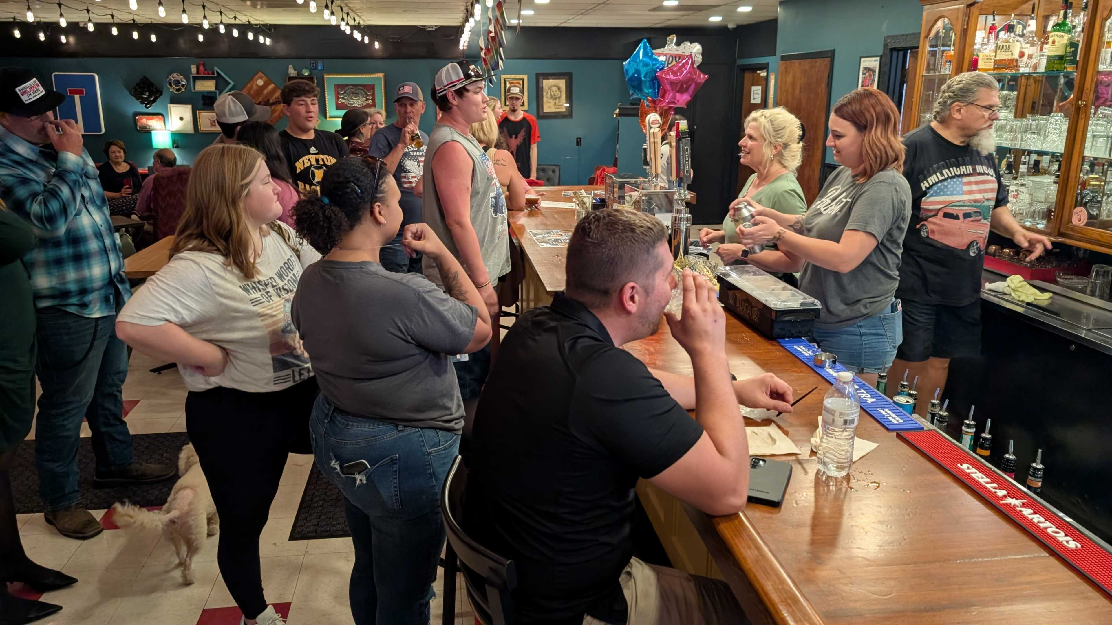 A diverse group of people gathers at a bar with a long counter, where bartenders serve drinks to customers while balloons and decorations adorn the space.