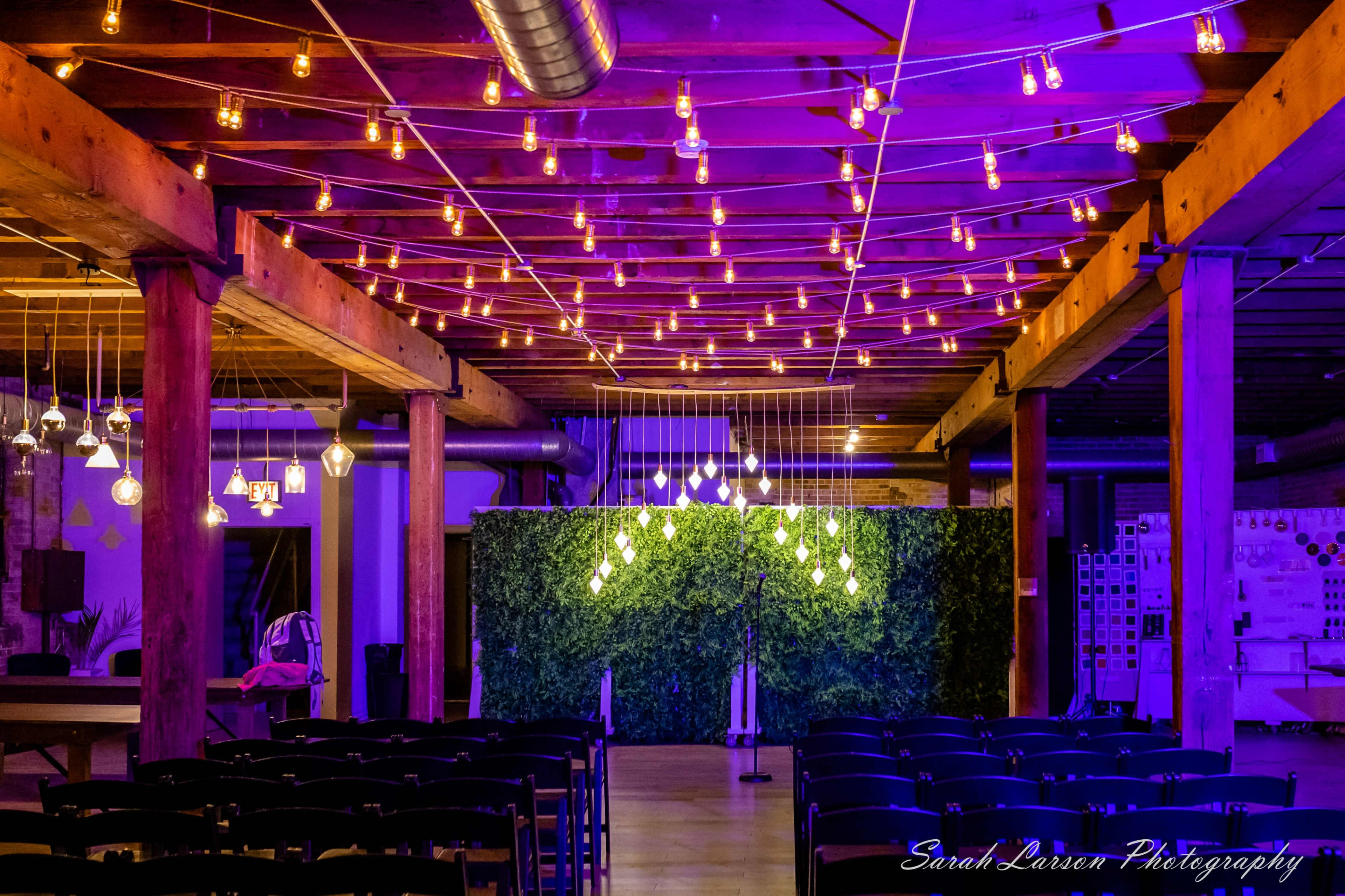 The image shows an event space with purple lighting and a ceiling adorned with hanging light bulbs, featuring a green backdrop and rows of chairs set up for guests.