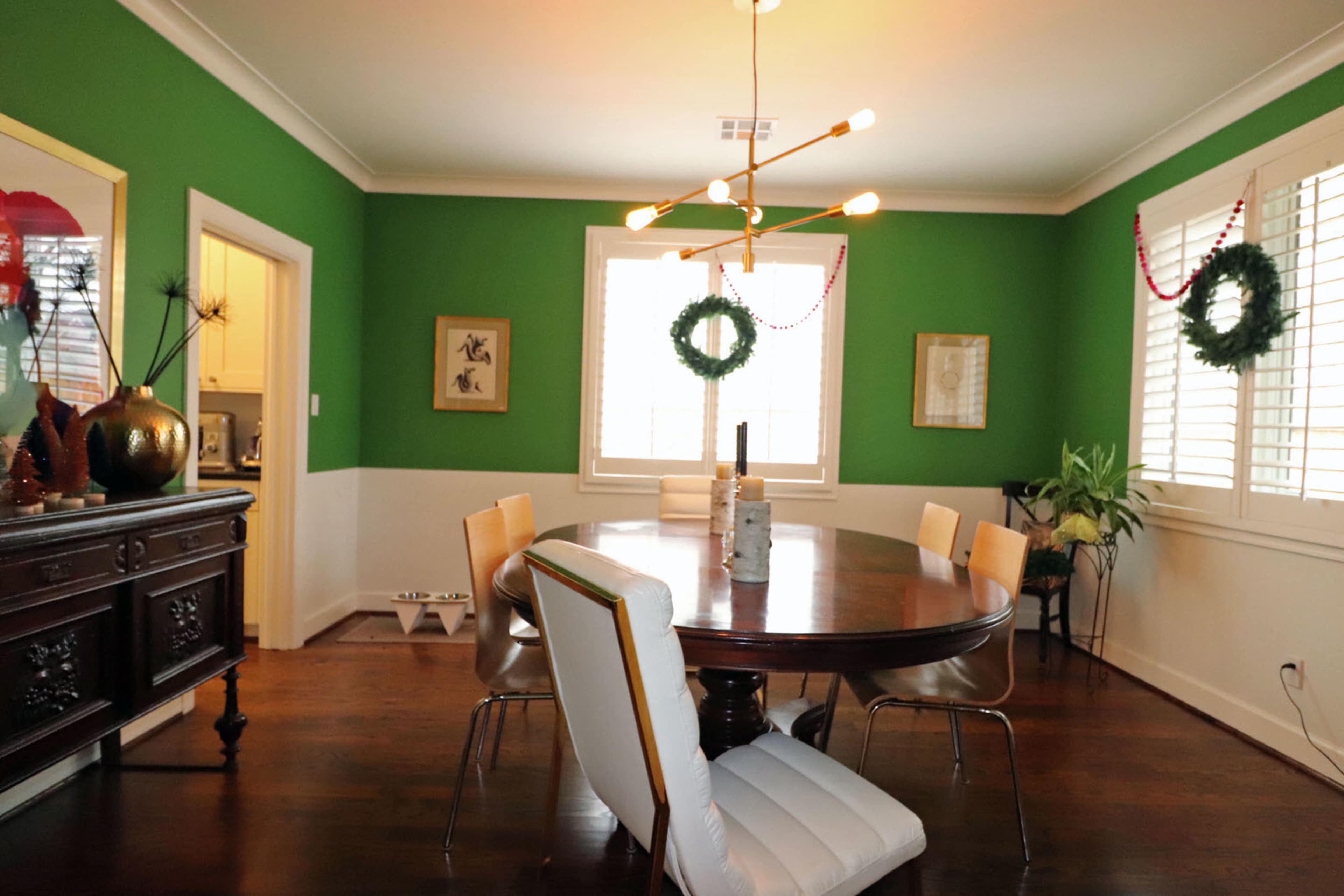 The dining room features a round wooden table surrounded by modern chairs, with green walls and festive decorations.